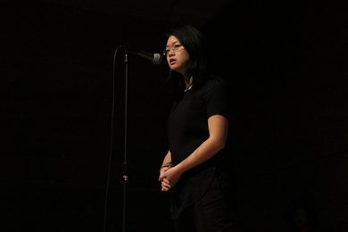 An Asian woman dressed in black speaks in front of a microphone.