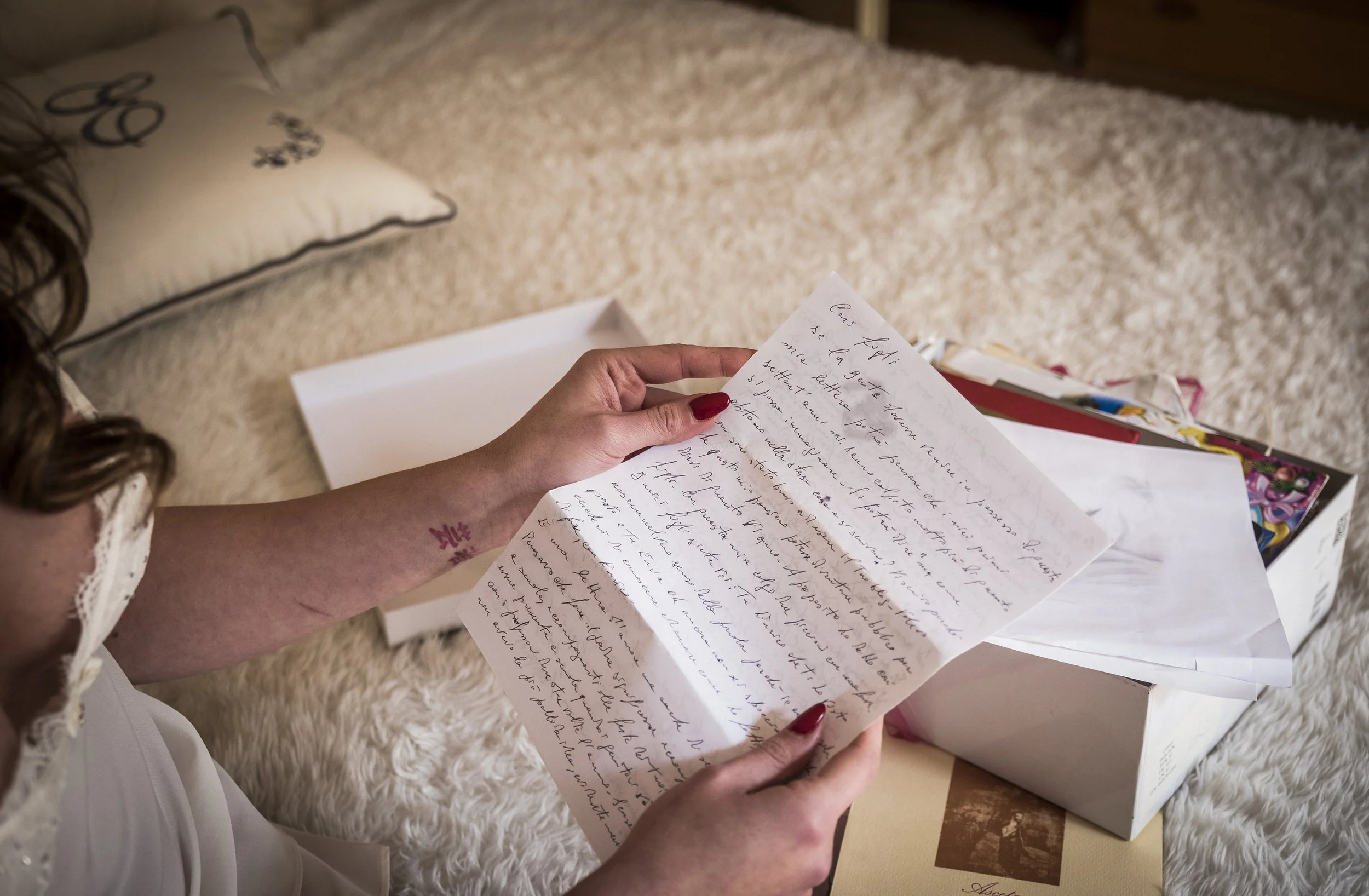 A woman with red nails and a floral tattoo on her wrist is holding a handwritten letter, sitting on a fluffy white rug with a pillow nearby and some colorful papers or postcards in the background.