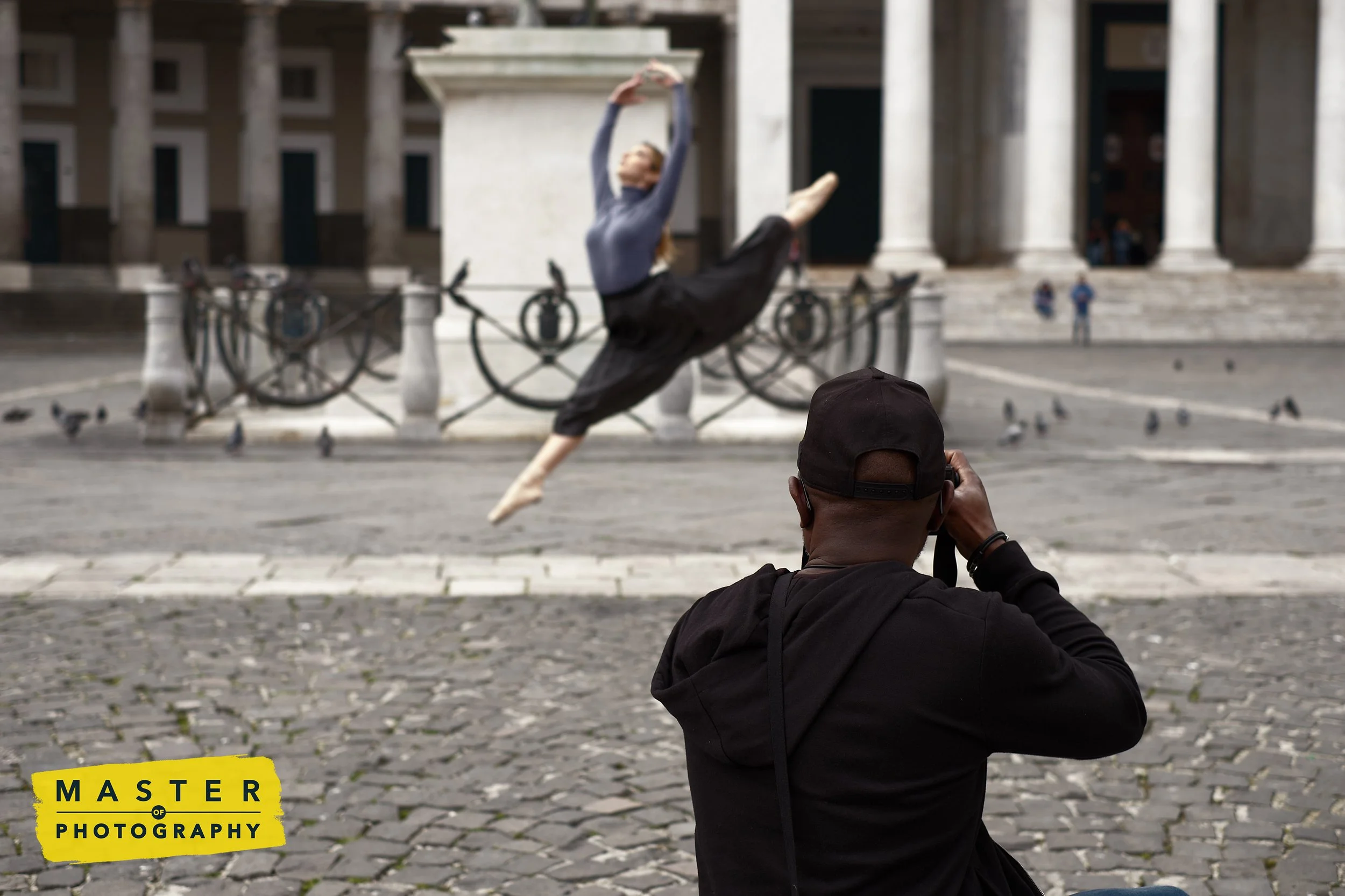 A man is taking a photo of a dancer performing a ballet pose in front of a large building with columns. The dancer is mid-air, with one leg extended behind and arms raised overhead. Several bicycles are parked behind a railing, and pigeons are scattered on the ground. The scene appears to be in a city square.