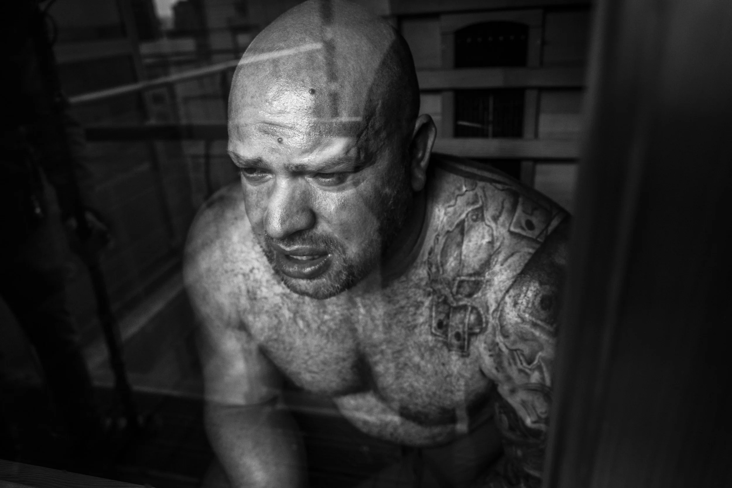 Black and white photo of a tattooed man with a bald head, looking downward. The scene appears to be inside or behind a metal fence or bars.