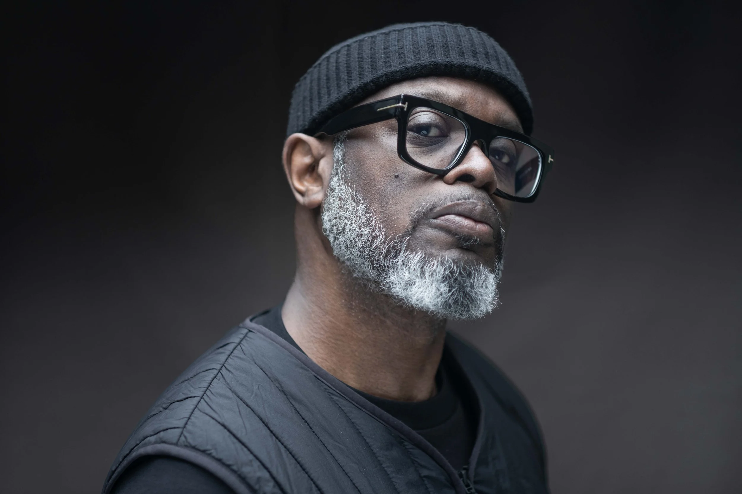 A close-up portrait of a middle-aged African American man with a gray beard wearing a gray beanie, black glasses, and a black quilted jacket against a dark background.