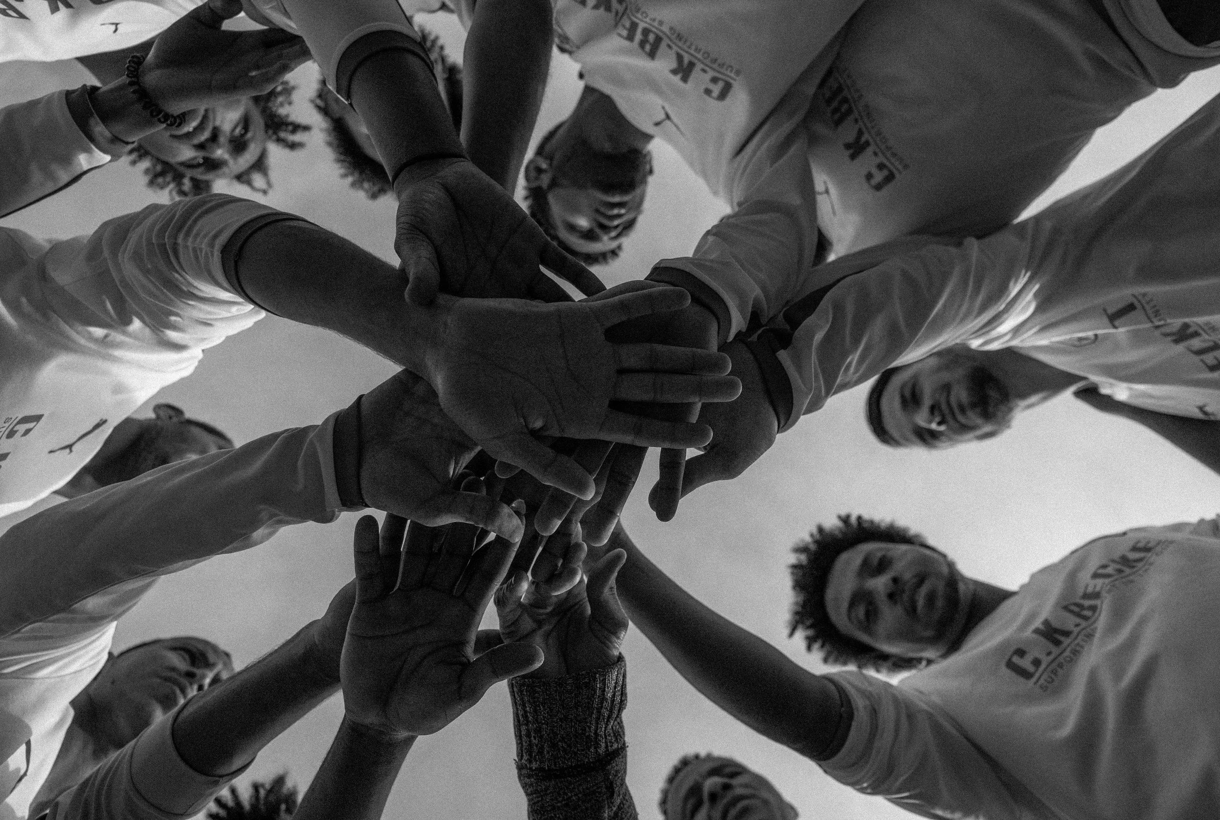 Group of people in sports jerseys huddling in a circle, placing their hands together for a team cheer, taken from below in black and white.