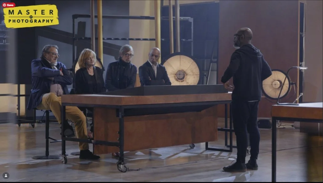A man stands in front of a panel of four people sitting at a table in a photography studio, with large studio lights and equipment behind them.