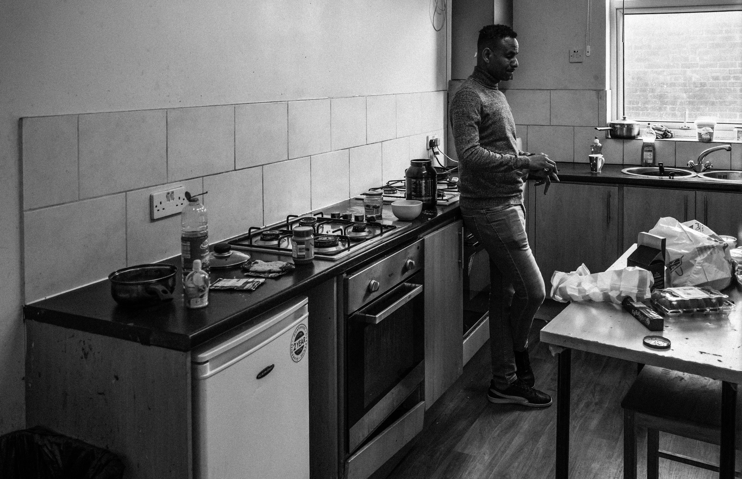 A man standing in a kitchen, looking at his phone. The kitchen has a gas stove, various food items, and kitchen utensils on the counters. The image is in black and white.