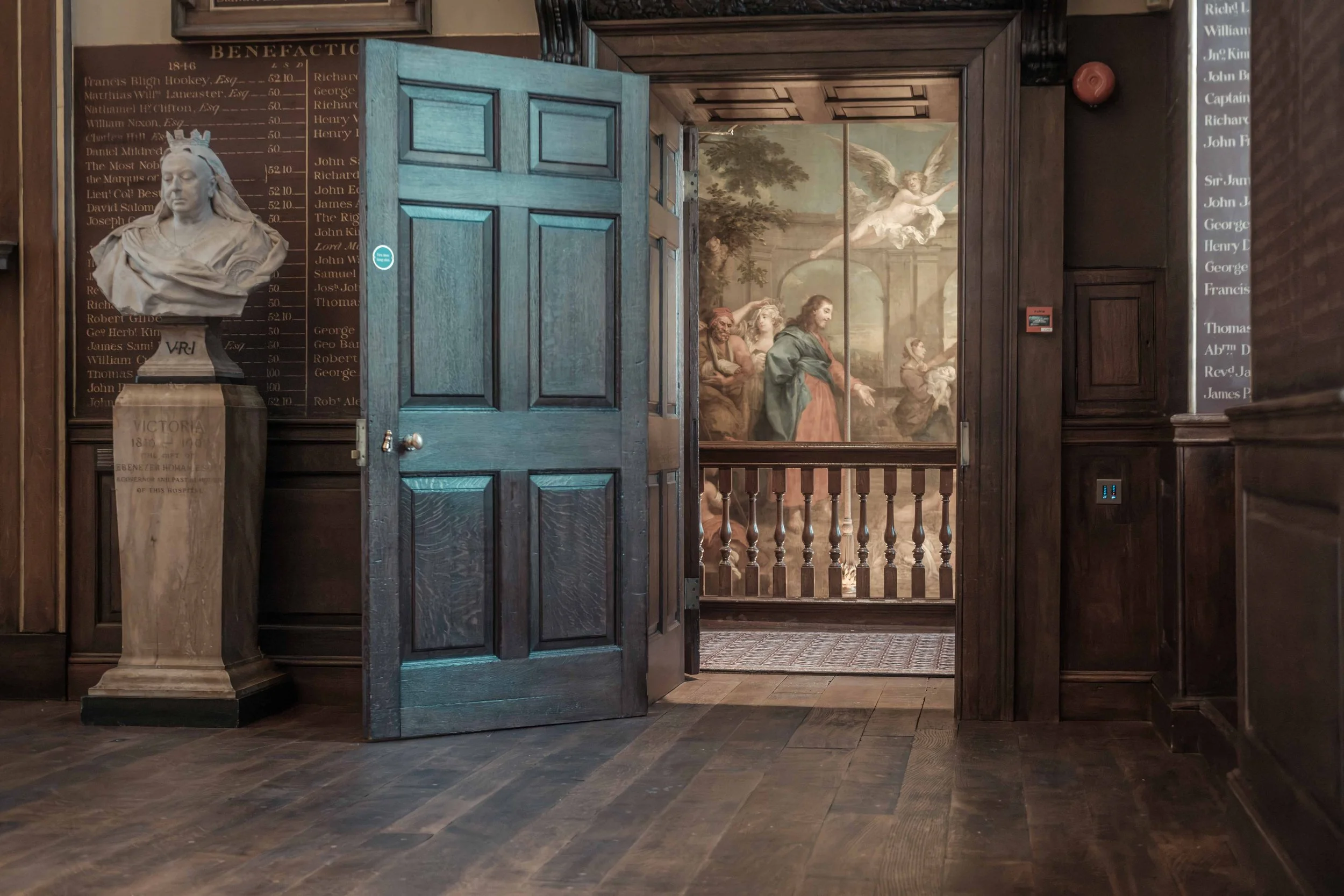 Open wooden door inside St Bartholomew's Hospital, revealing a marble bust, historic benefactors' boards and a painting beyond.