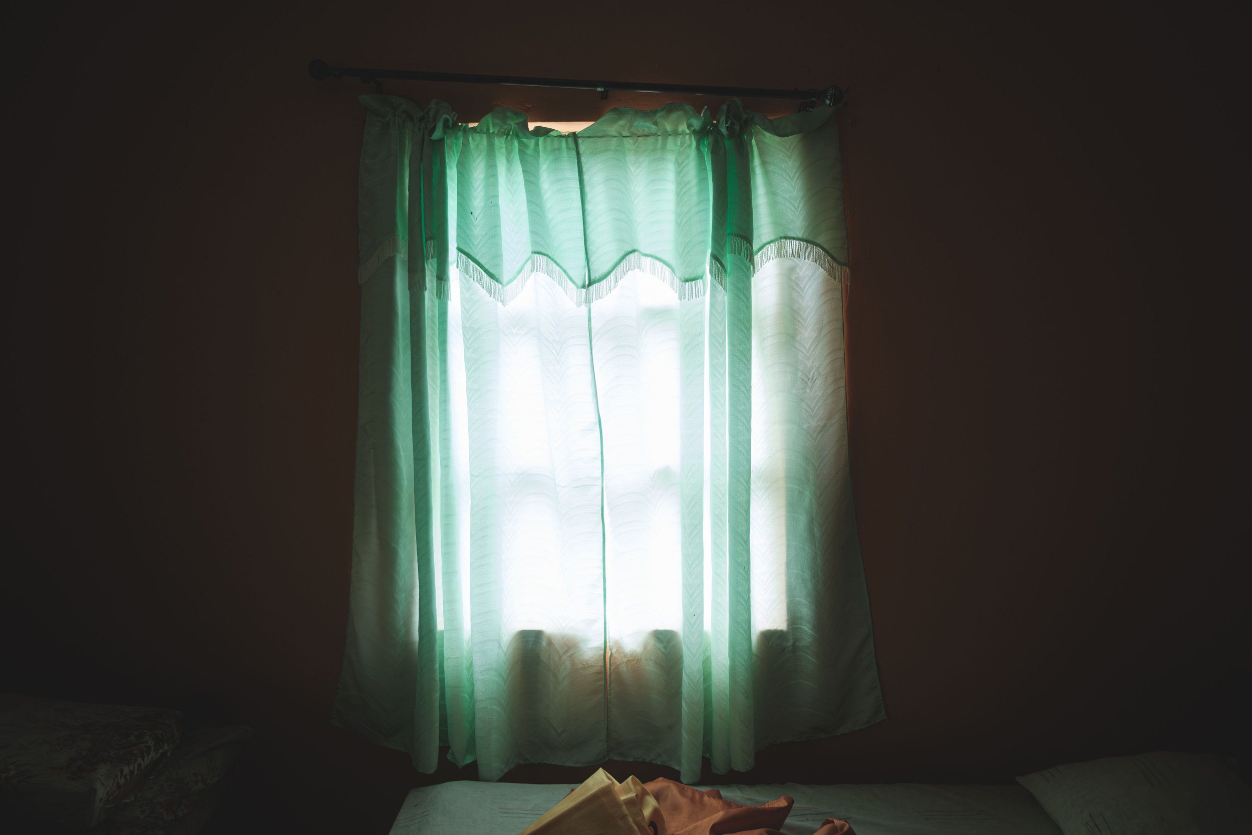 Green curtains hanging over a window with light shining through, casting shadows inside a dim room.