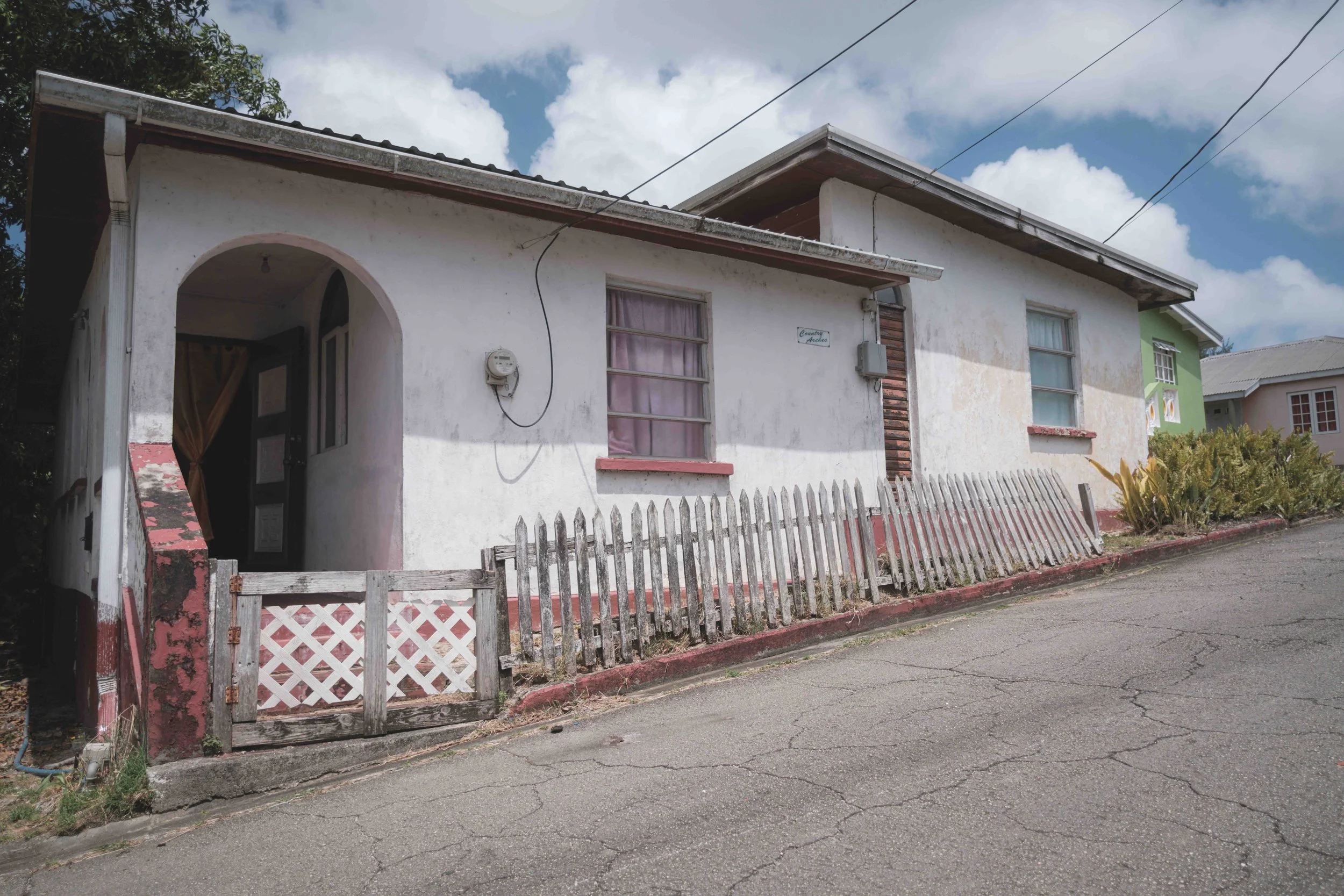 A small, weathered house with white walls, pink trim, and a wooden fence along the sidewalk. There are two windows with curtains, a door, and an overgrown yard. The house is located on a street with visible cracks, and the sky is partly cloudy.