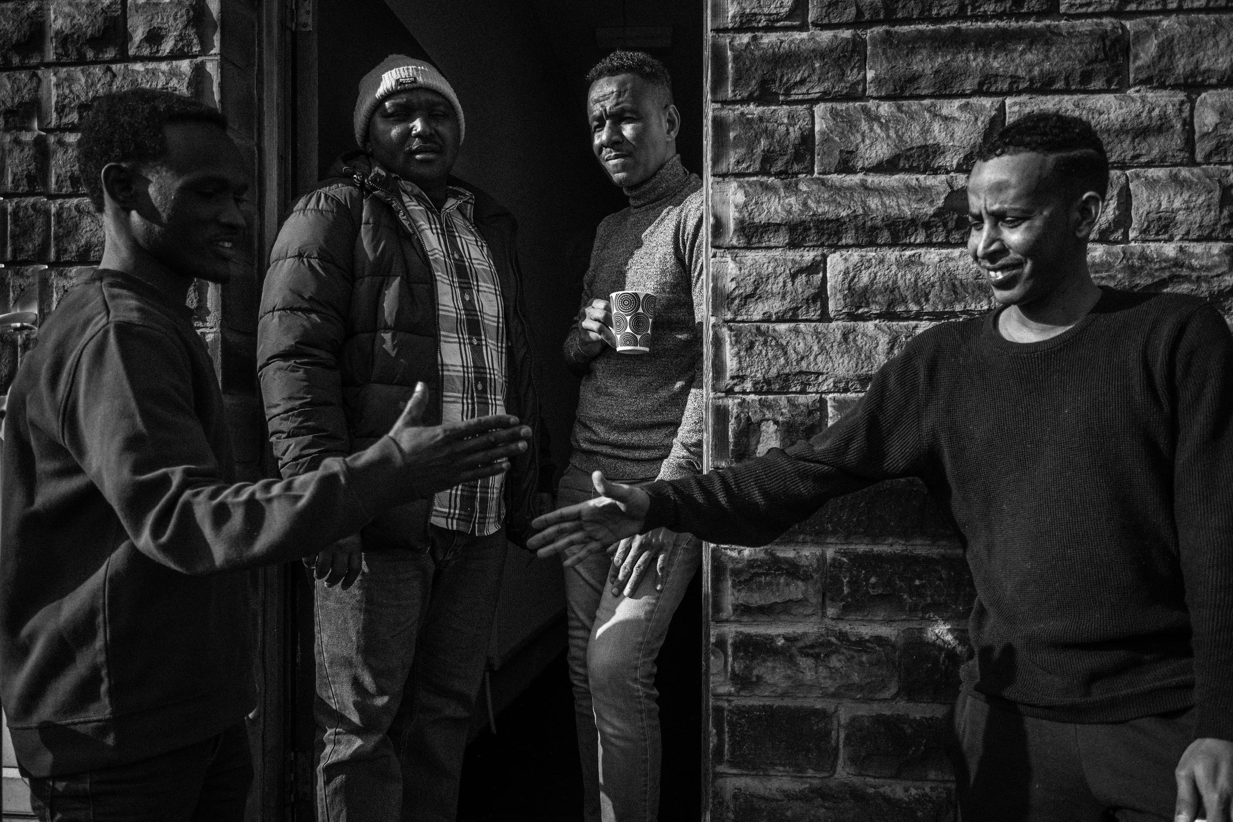 Four young African American people standing and talking outside, near a brick wall, with some smiling and gesturing.