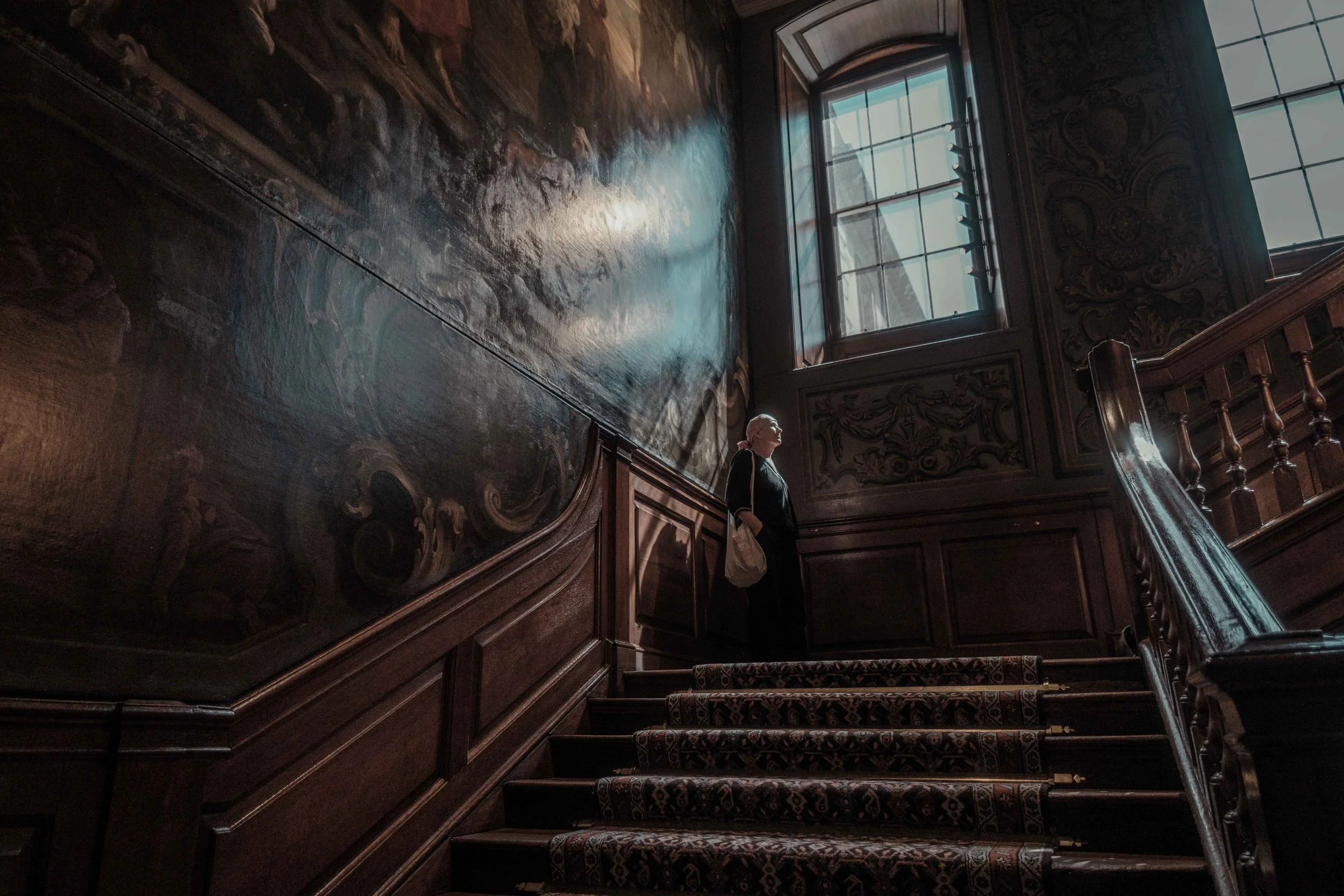 Visitor standing on the historic Hogarth Staircase at St Bartholomew's Hospital, surrounded by dark wooden panelling and paintings illuminated by natural light.