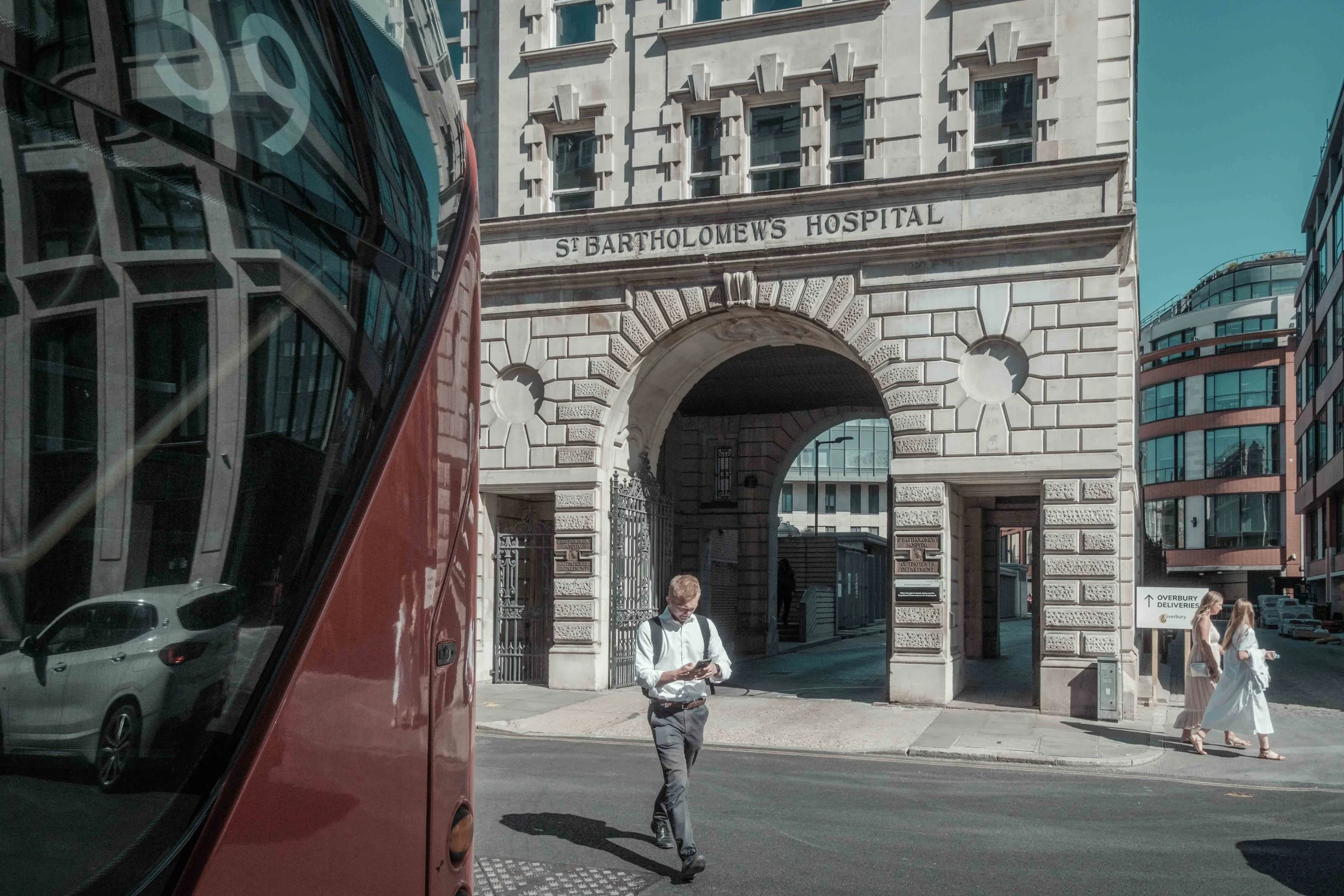 Red London bus passing the historic stone entrance to St Bartholomew's Hospital in London, with pedestrians crossing the street.