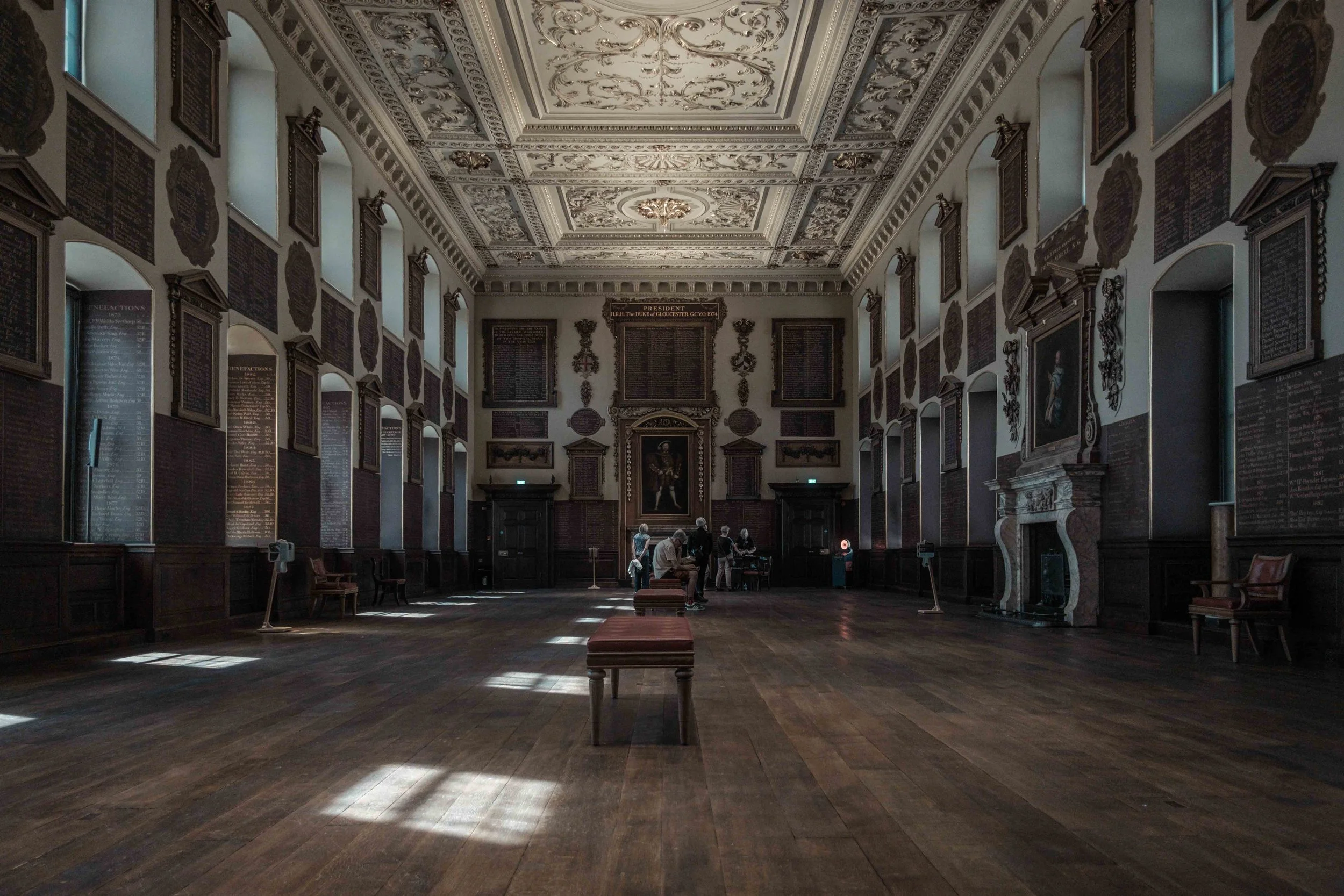 Wide interior view of the Great Hall at St Bartholomew's Hospital, showing its decorated ceiling, wooden floor and historic benefactors' boards.