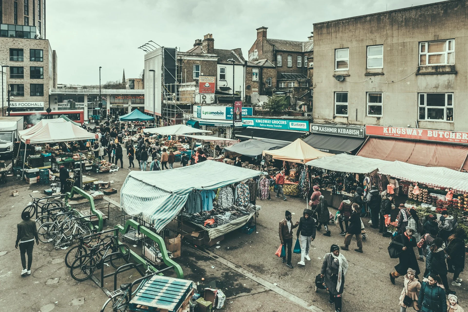 Street market scene with tents and vendors selling goods, people walking, bicycles parked, and buildings in the background.