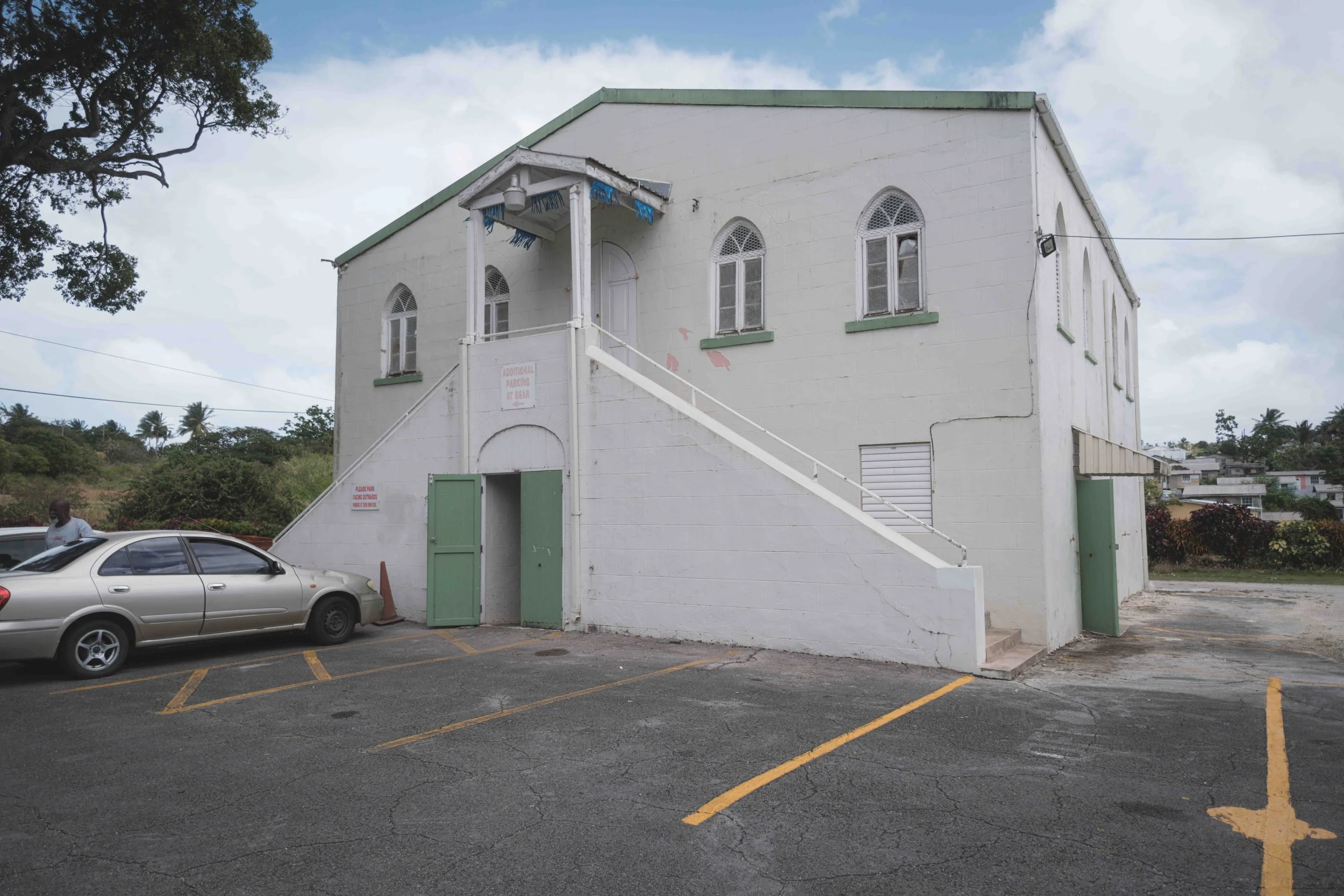 White two-story building with green trim and windows, a staircase with a green door at the base, parking lot with cars, and a tree to the left with cloudy sky overhead.