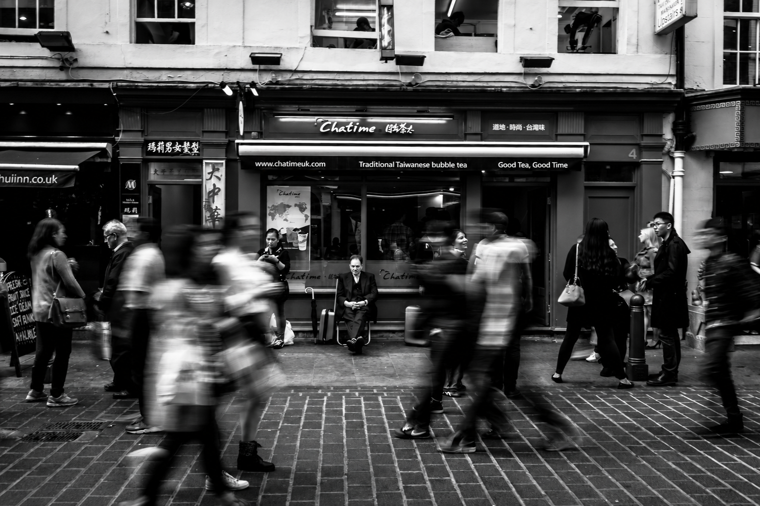 Black and white photo of a busy street scene in front of a tea shop called 'Chatime'. People are walking past, some blurred by motion, and a man waits alone seated outside with a suitcase. Inside the shop, people are visible through the window, engaged in activities.