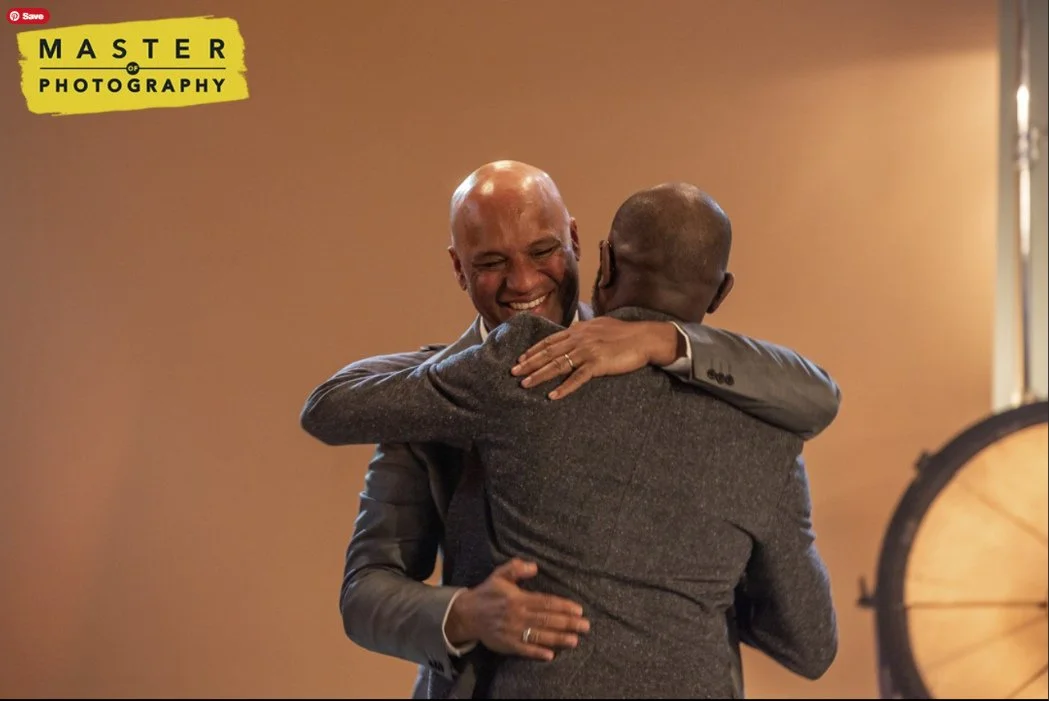 Two men with gray suits hugging and smiling in a warm setting.
