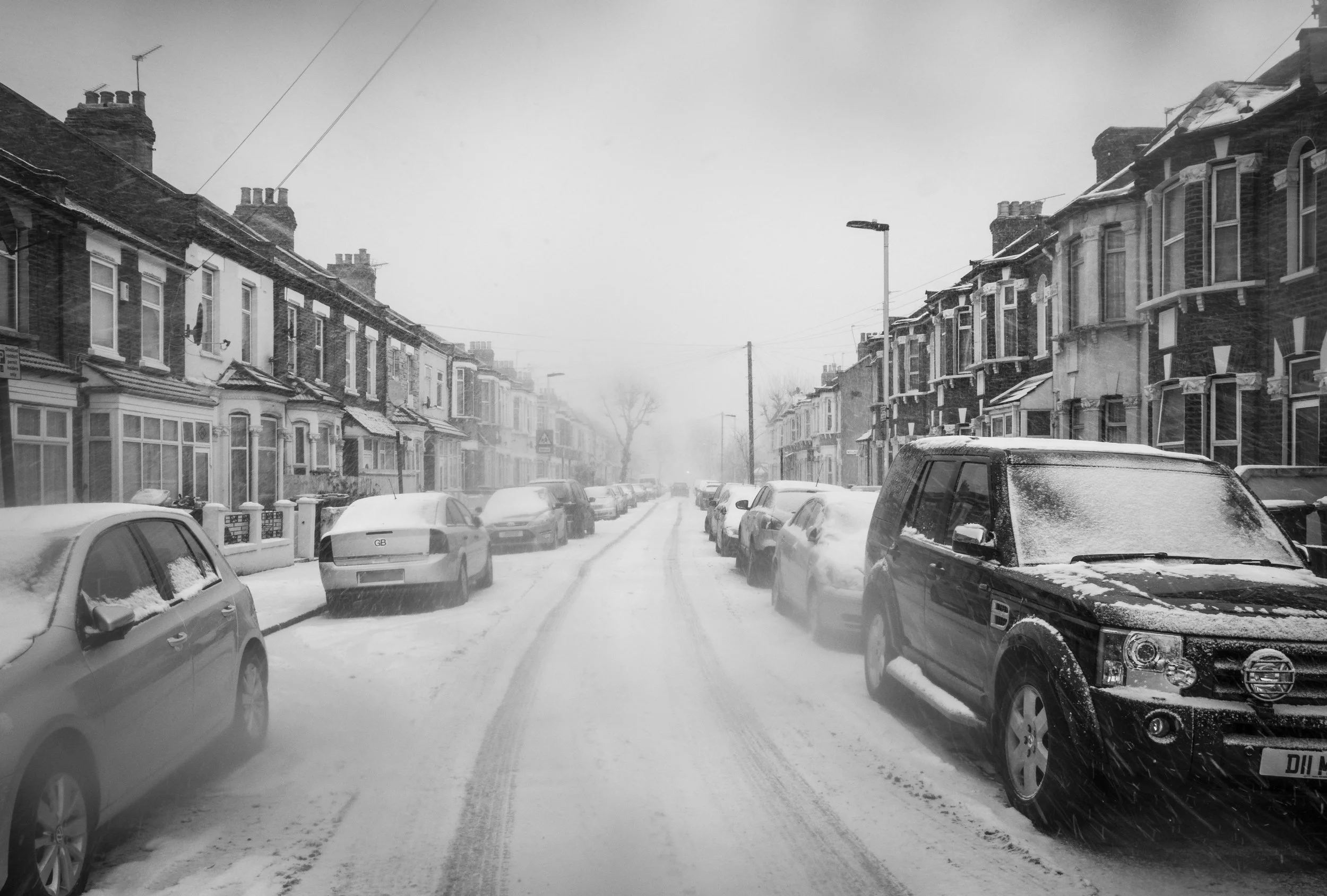 Snow-covered residential street with parked cars and row of brick houses, some with bay windows, under cloudy sky.