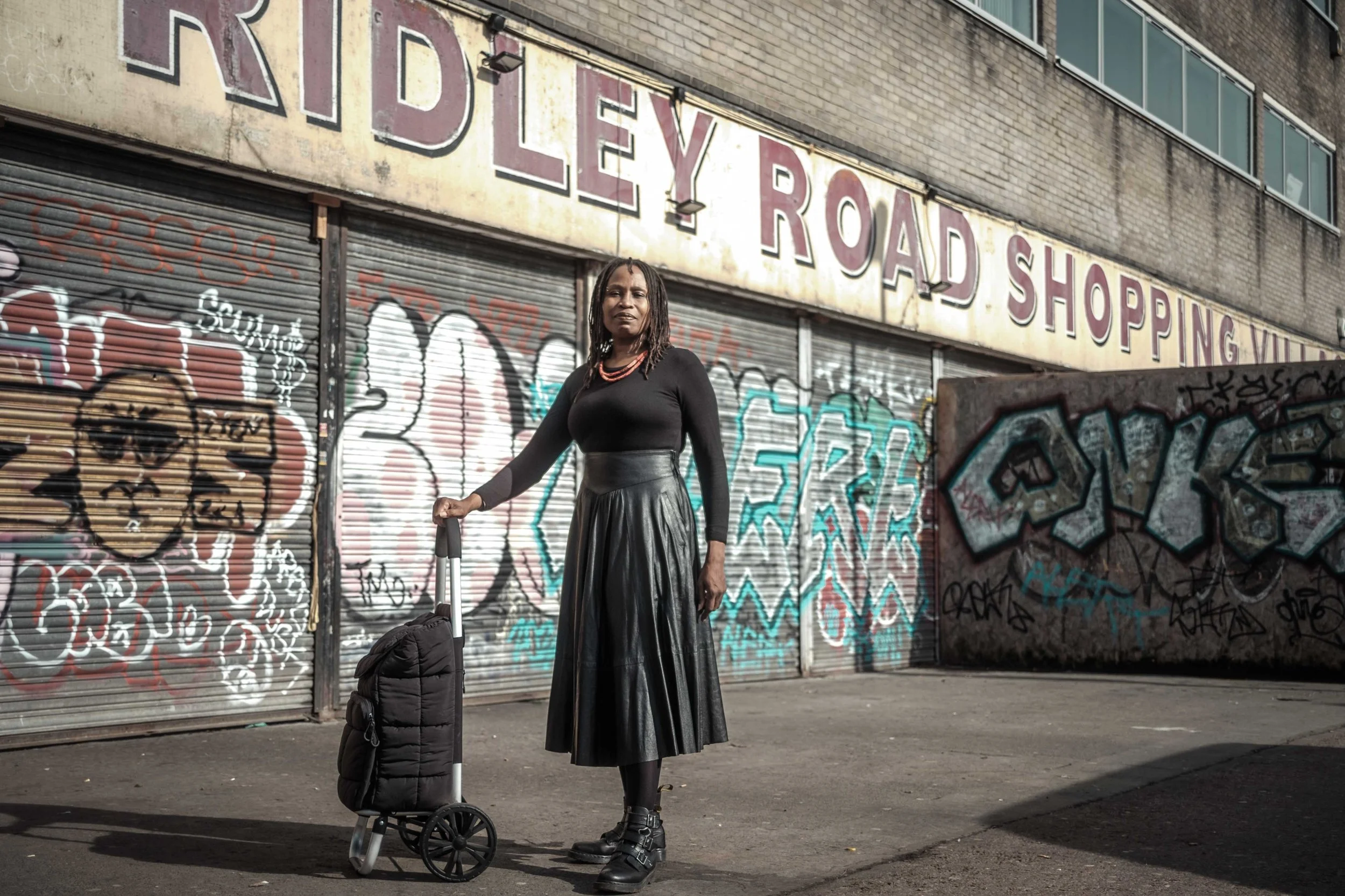 A woman with dreadlocks wearing a black long-sleeve top and a long skirt standing in front of a graffitied storefront with a closed metal shutter, holding a black rolling suitcase, with a black coat draped over a trolley