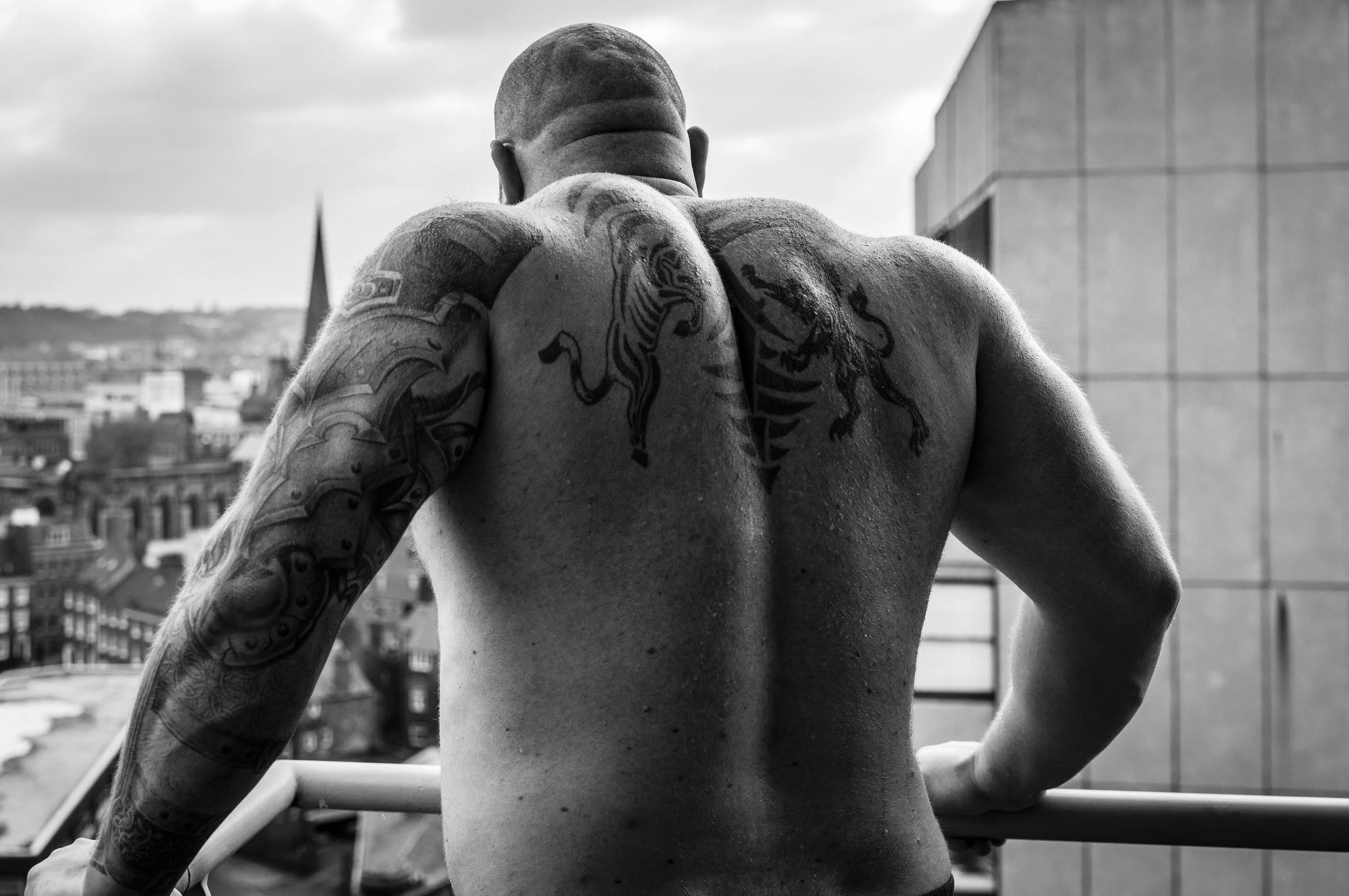 Back of a shirtless man with tattoos on his arms and back, looking out over city rooftops.