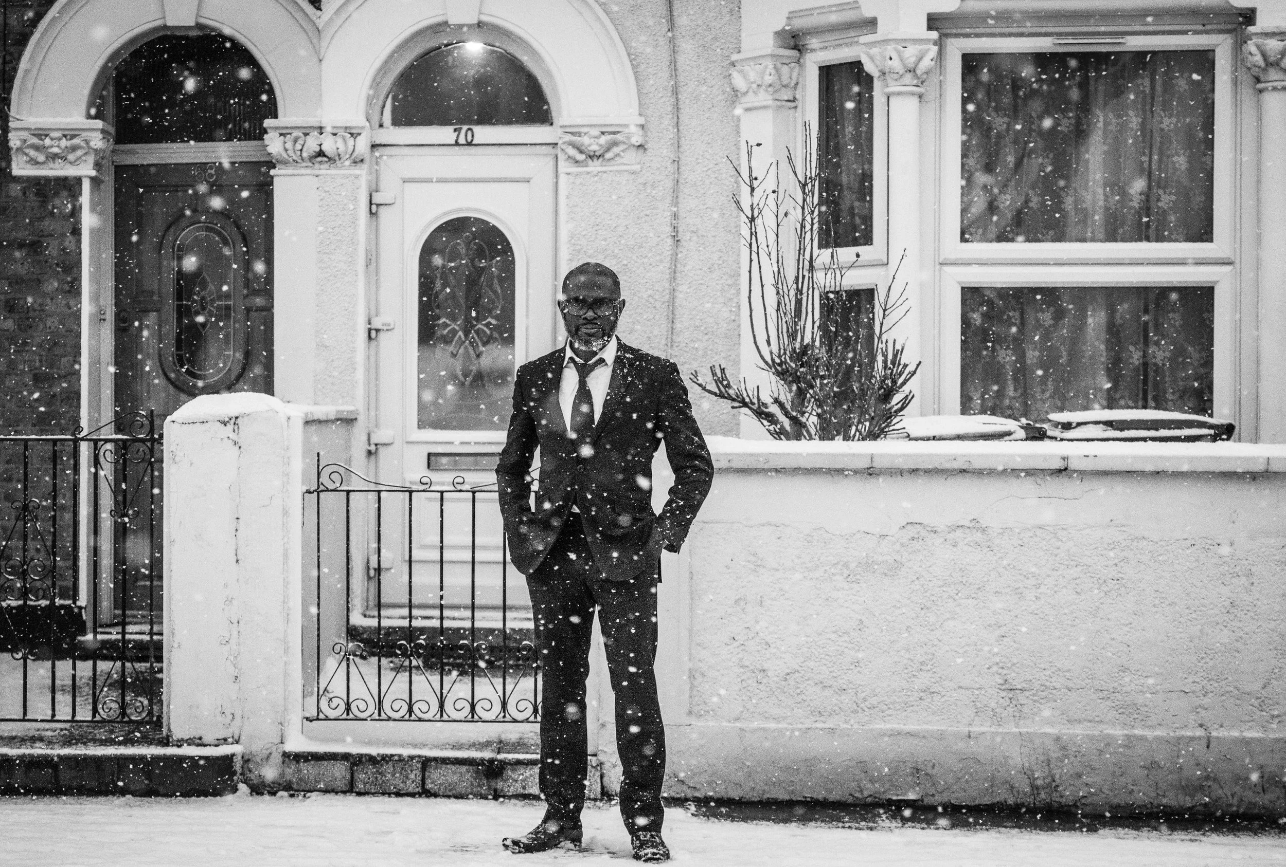 A man in a suit with a tie and glasses standing outside in front of a house during snowfall. The house has a decorative door with a glass window and arched window above, along with a large window to the right and a small bush in the front yard.