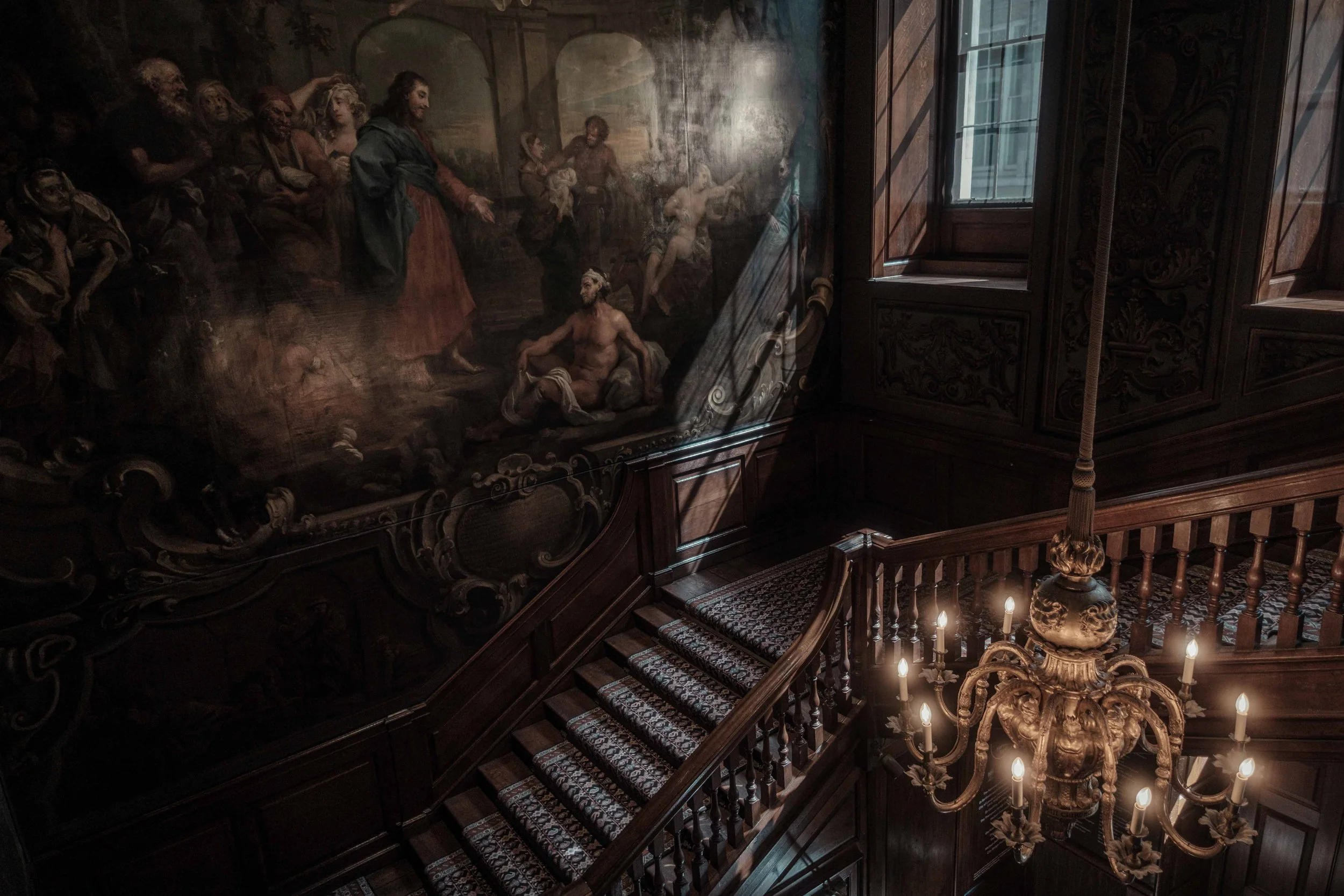 Ornate wooden staircase at St Bartholomew's Hospital, with a large painting by William Hogarth and an illuminated chandelier.