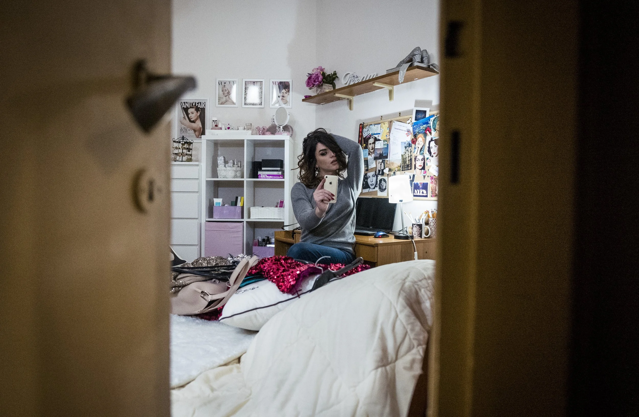 A woman taking a selfie in her bedroom, sitting on a bed surrounded by clothes and accessories, with decorative items and a bulletin board on the wall behind her.