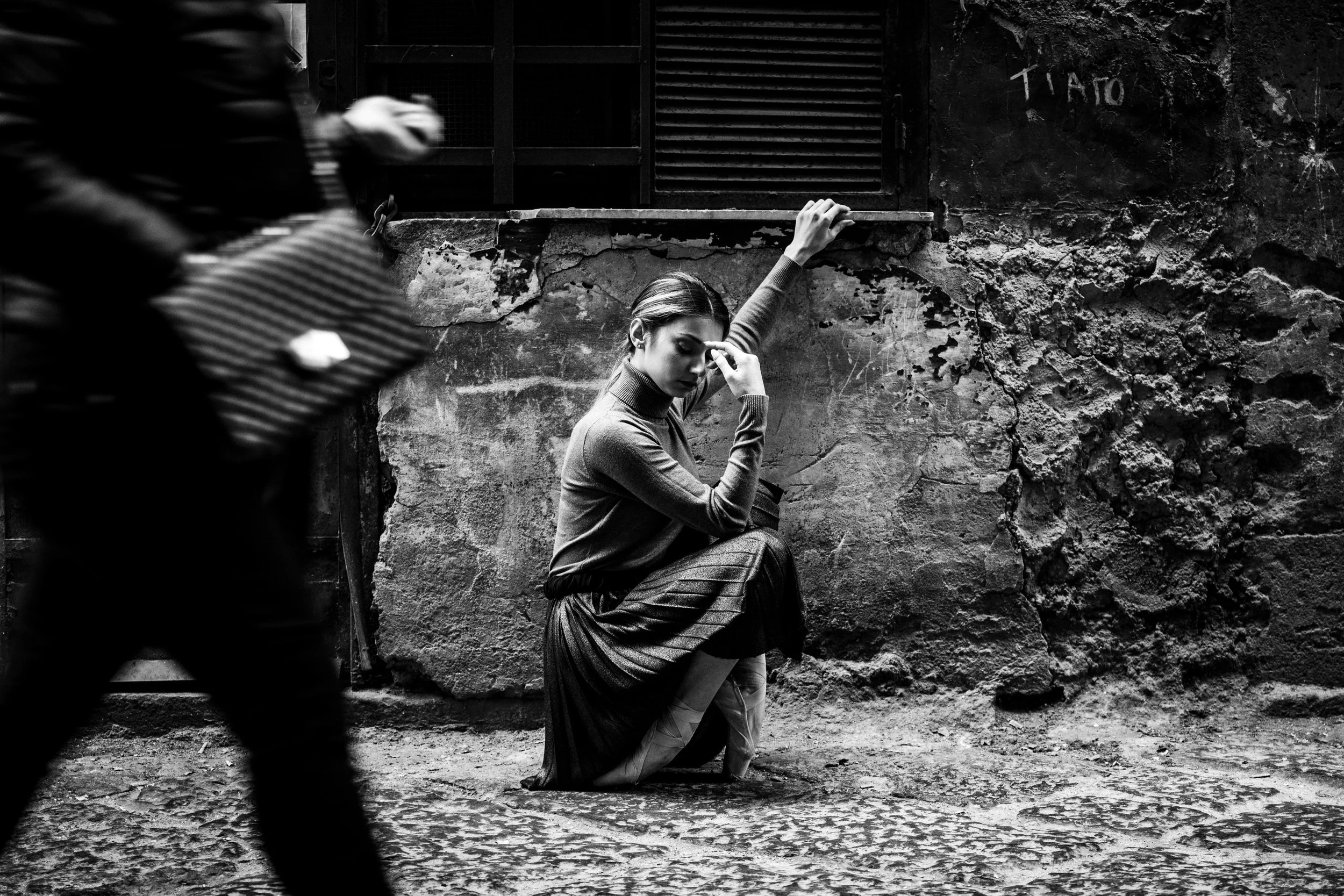 A woman with short hair, dressed in a long skirt and sweater, is sitting on the ground with her eyes closed and a hand resting on her forehead, leaning against a textured, weathered wall in an alleyway. A blurred figure is passing by in the foreground. The scene is in black and white.