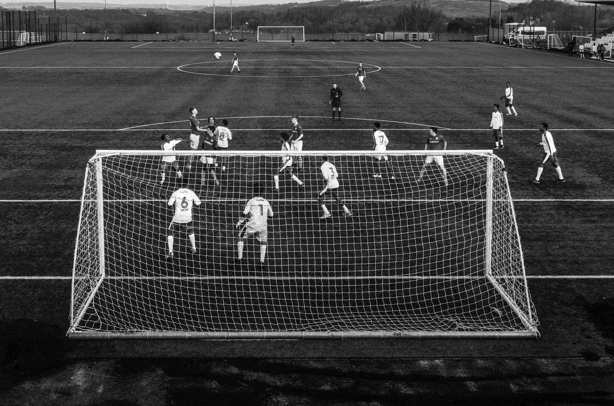 Black and white photo of a soccer game on a field, with players near the goal and others on the field, with a goal net in the foreground.