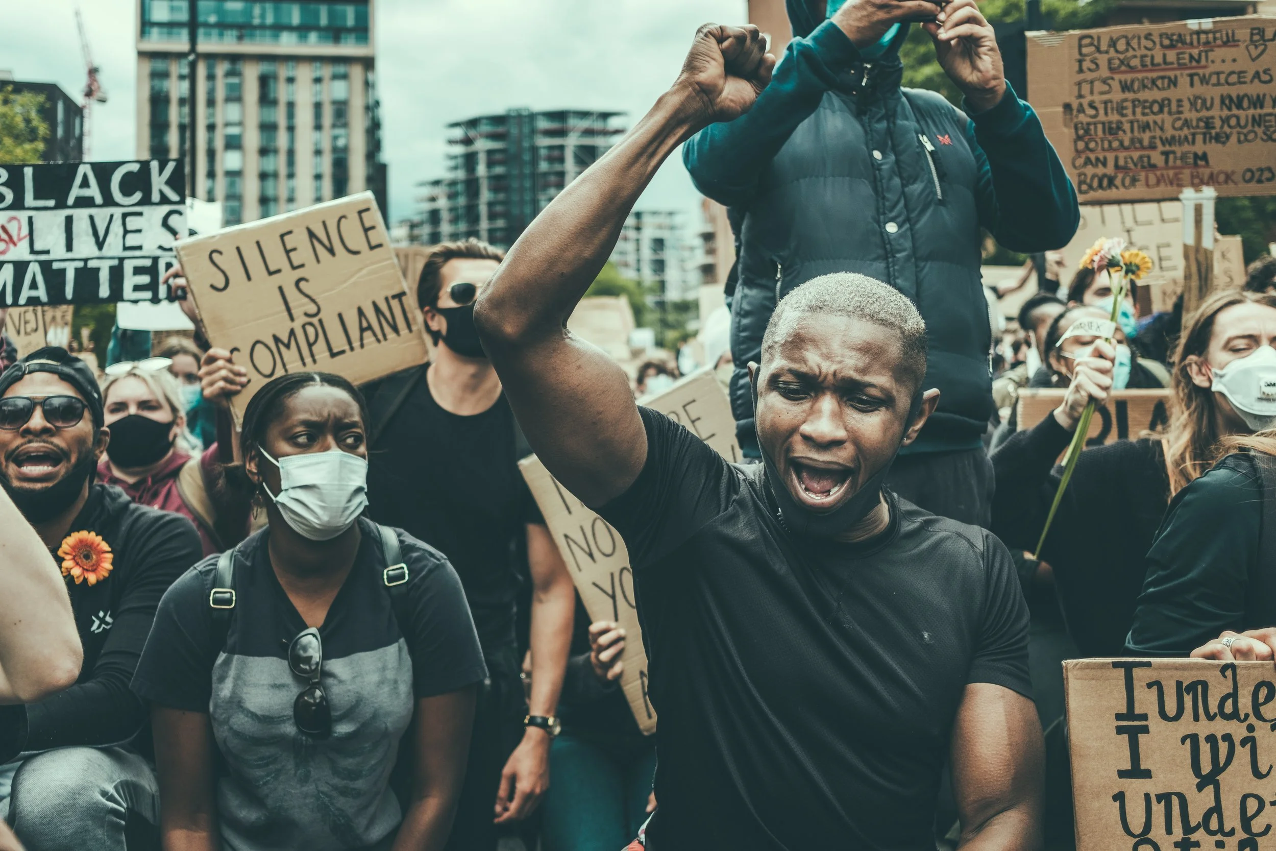 A diverse group of protesters, including men and women wearing masks, participating in a rally. They are holding various signs, including one that reads 'SILENCE IS COMPLIANT' and another that says 'BLACK LIVES MATTER.' The crowd appears to be outdoors in an urban area with high-rise buildings visible in the background.