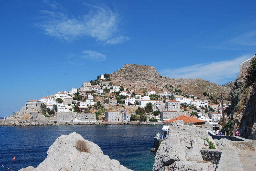 Rounding the corner into Hydra Port on the coastal path. Omilos Restaurant can be seen to the left of the path, down by the water. 