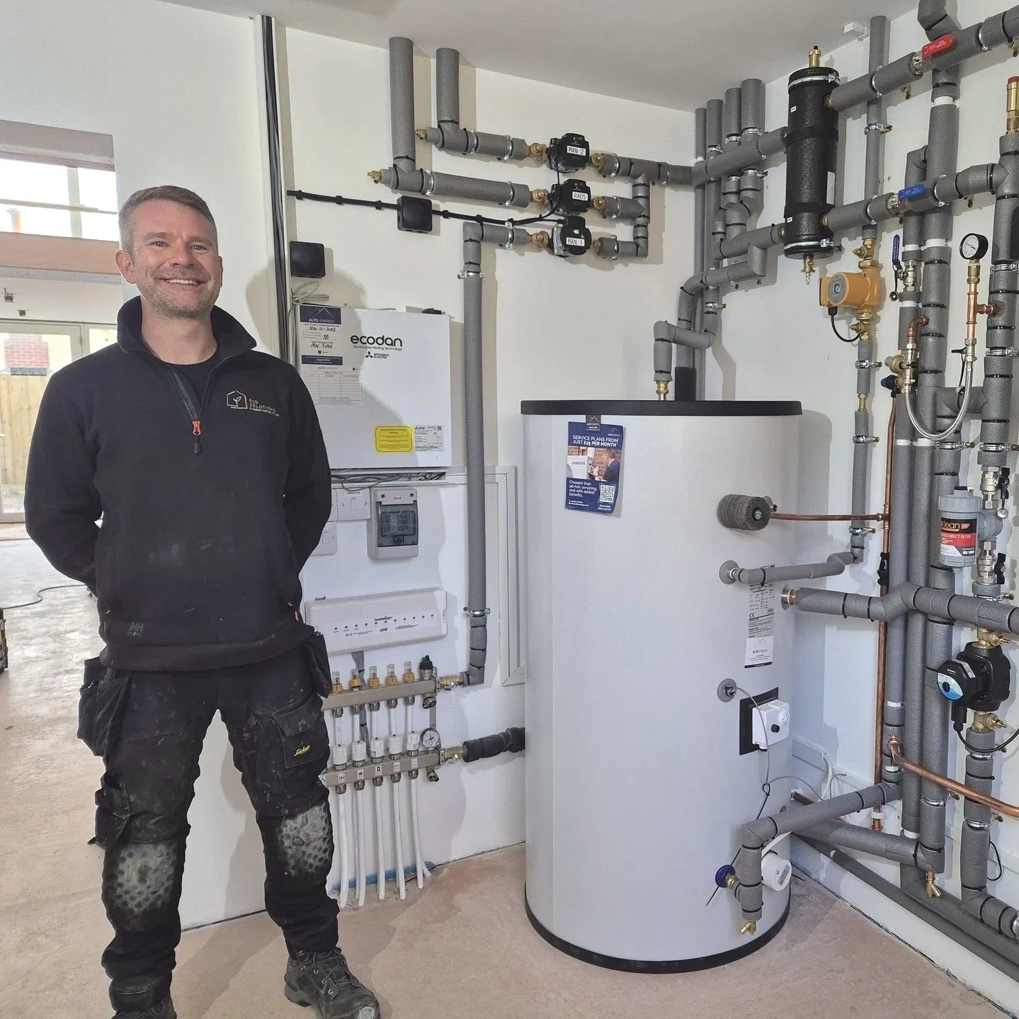 Partner installer from Eco Solutions Plumbing standing beside a completed Mitsubishi Ecodan heat pump installation, showing the indoor hot water cylinder and pipework.