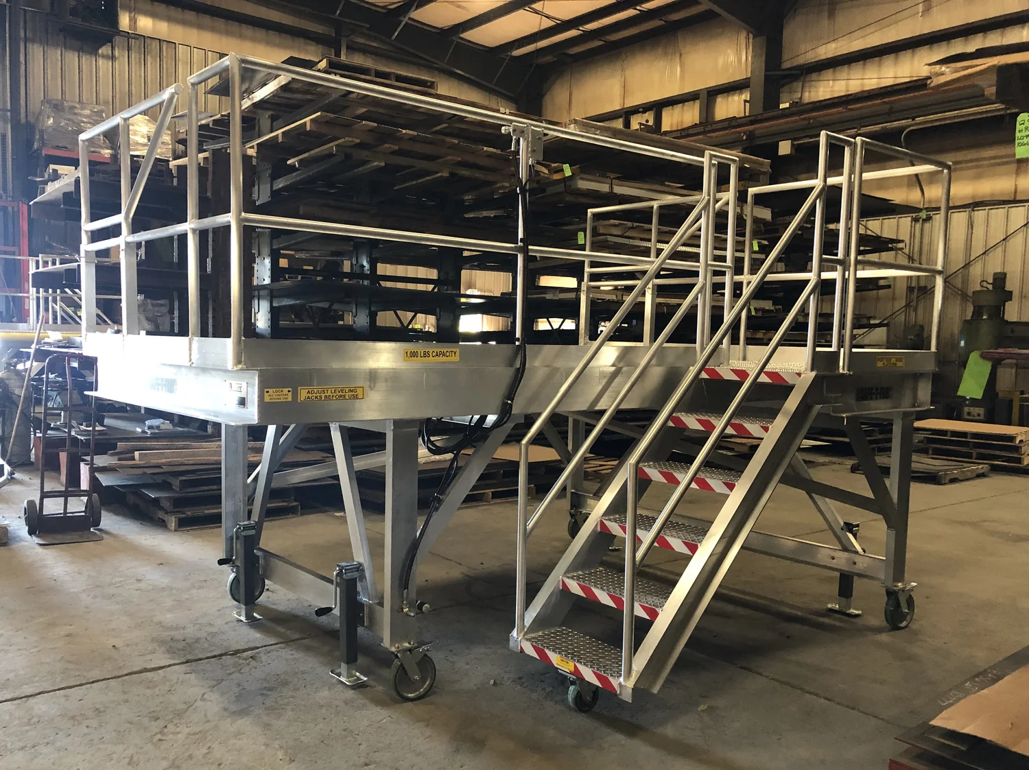 An aluminium SAFE-T-FAB mobile work platform on the shop floor — twin guarded decks reached by a stair with red-and-white hazard nosing, riding on locking casters with screw-down leveling jacks, rated 1,000 lb capacity.