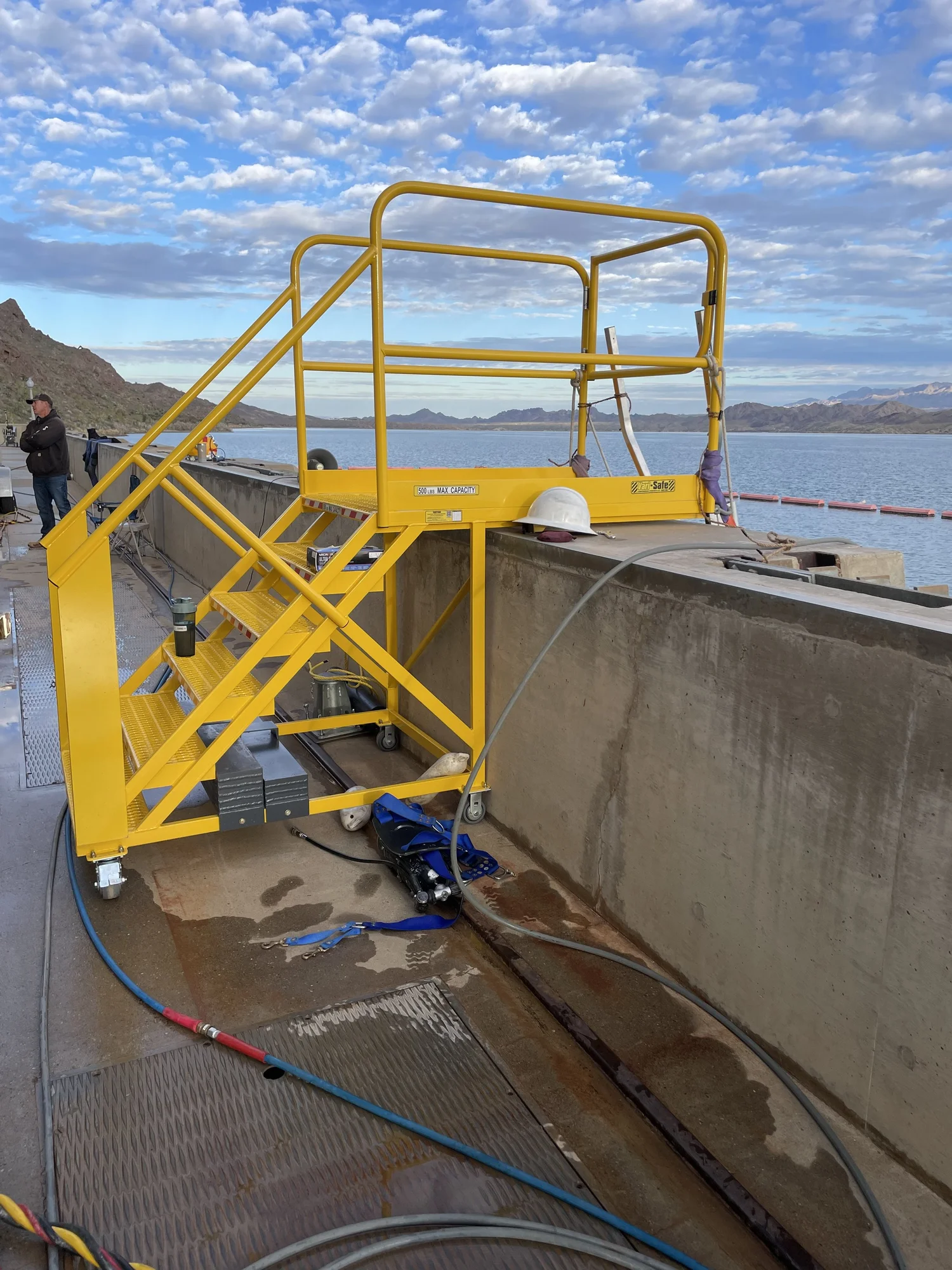 A yellow SAFE-T-FAB cantilever work platform reaching out over a concrete channel on an outdoor site, its counterweighted base standing clear of the edge.