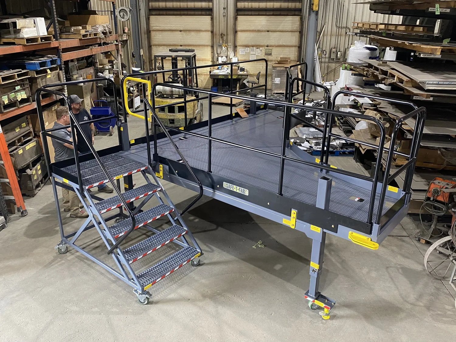 A SAFE-T-FAB mobile work platform on the shop floor — a wide diamond-plate deck inside black guardrails, reached by a stair with grip-strut treads and red-and-white hazard nosing, with two of the build team standing alongside for scale.