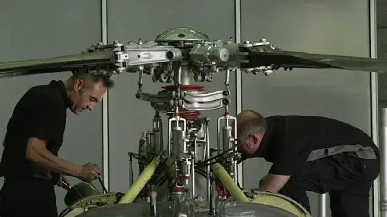 Two technicians working on the rotor head of a helicopter at hangar-floor level — the kind of overhead rotor access SAFE-T-FAB builds maintenance stands for.