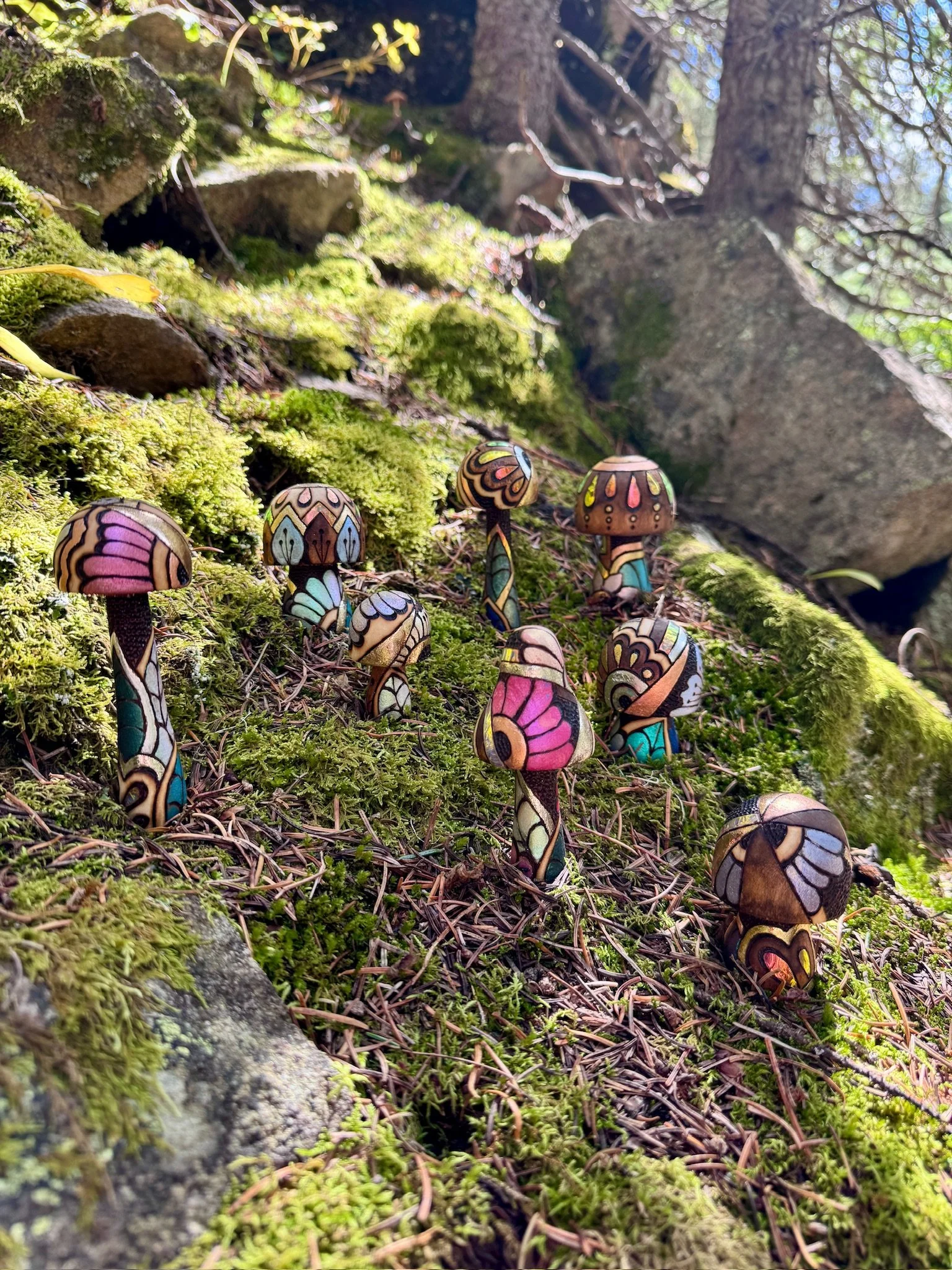 Light-responsive wooden mushroom sculptures with pink, turquoise, and metallic detailing displayed alongside wild mushrooms on a forest floor.