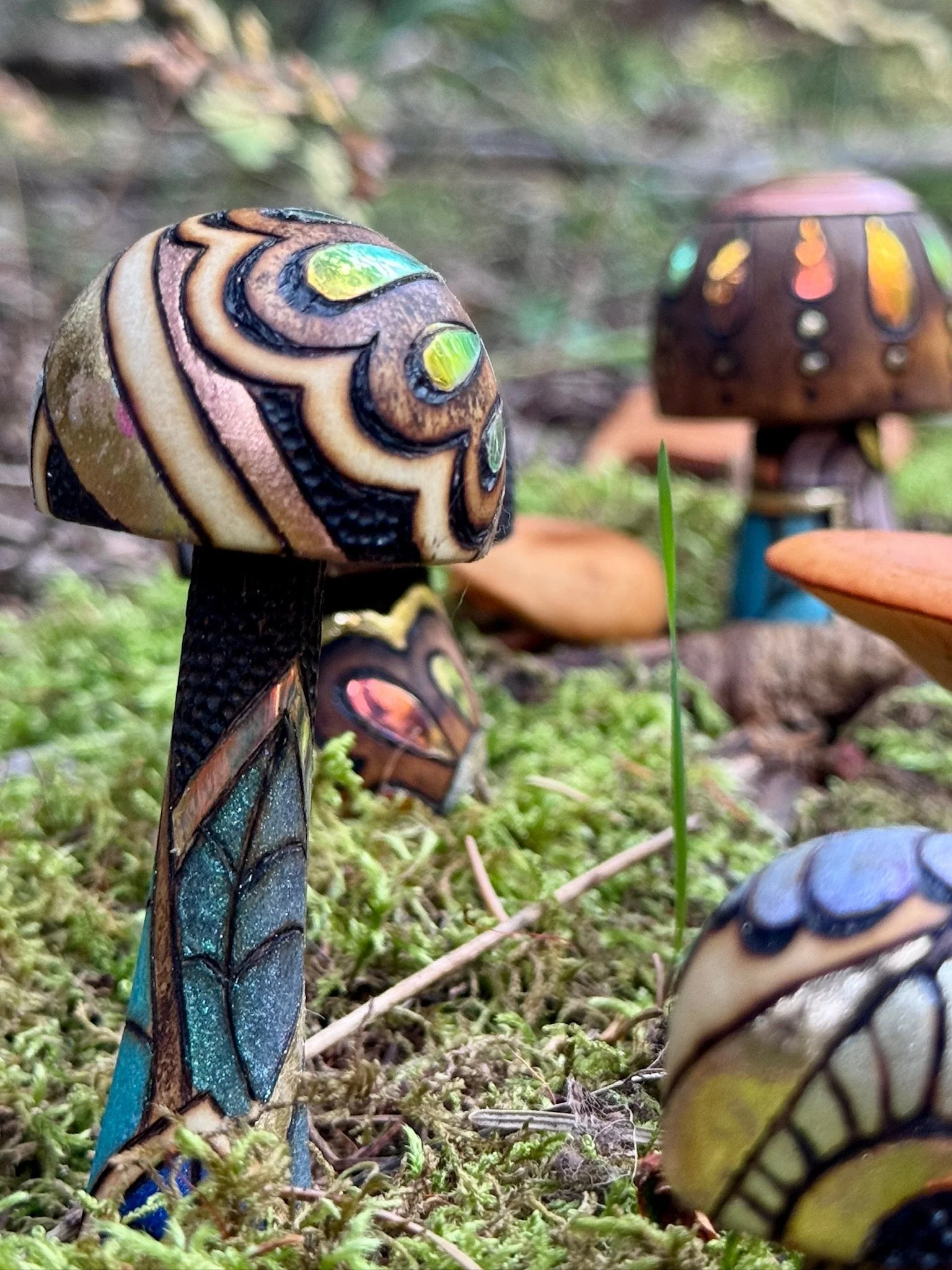 Close view of two colorful wooden mushroom sculptures with pyrography, gilded accents, and reflective patterns among moss and natural mushrooms.