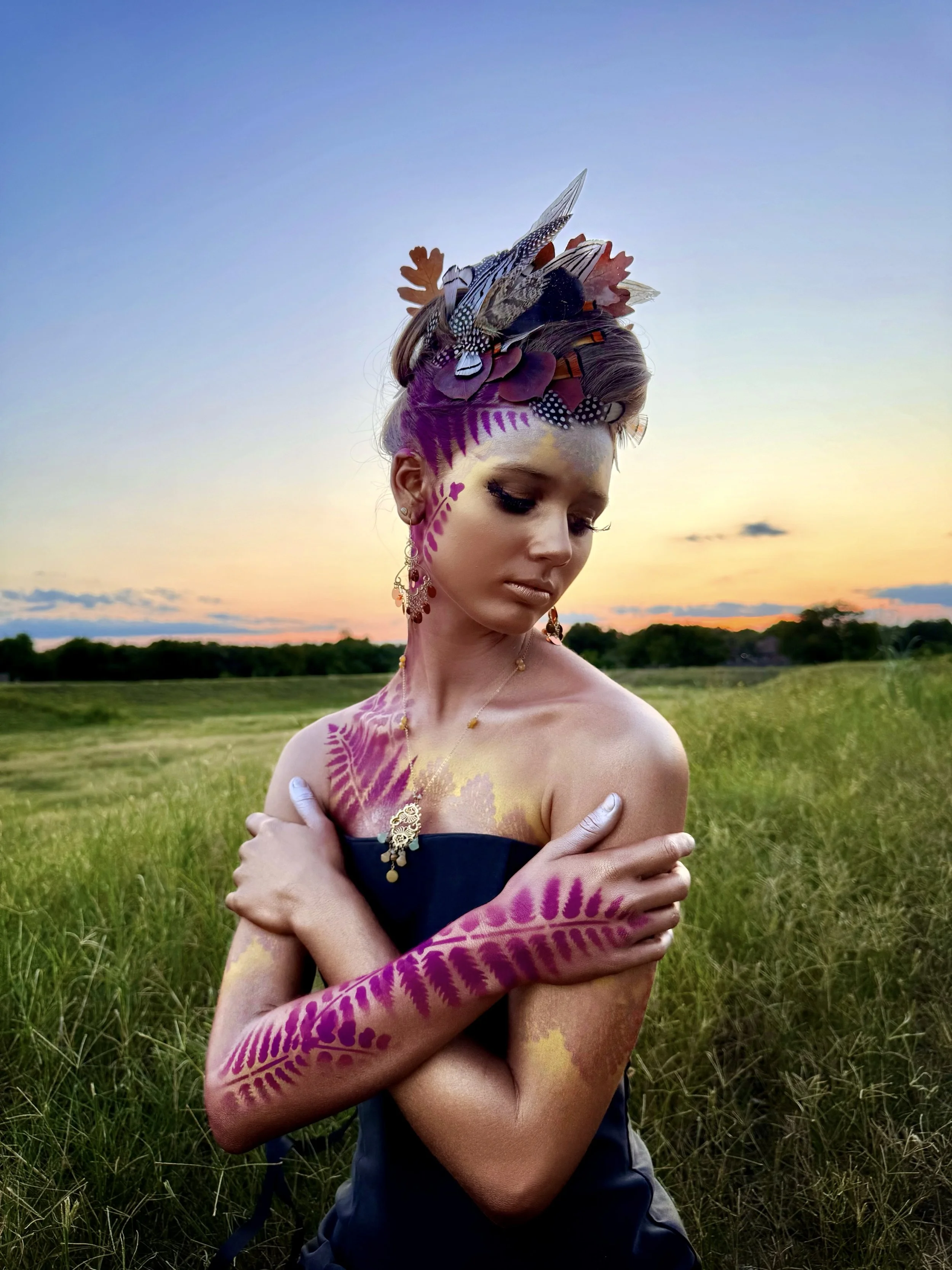  Golden hour in Austin, Texas frames this serene body art portrait by Anne Shackelford. The model is adorned in gold shimmer and magenta fern leaf patterns, wearing a custom feathered headpiece. The composition blends fashion photography with fine ar