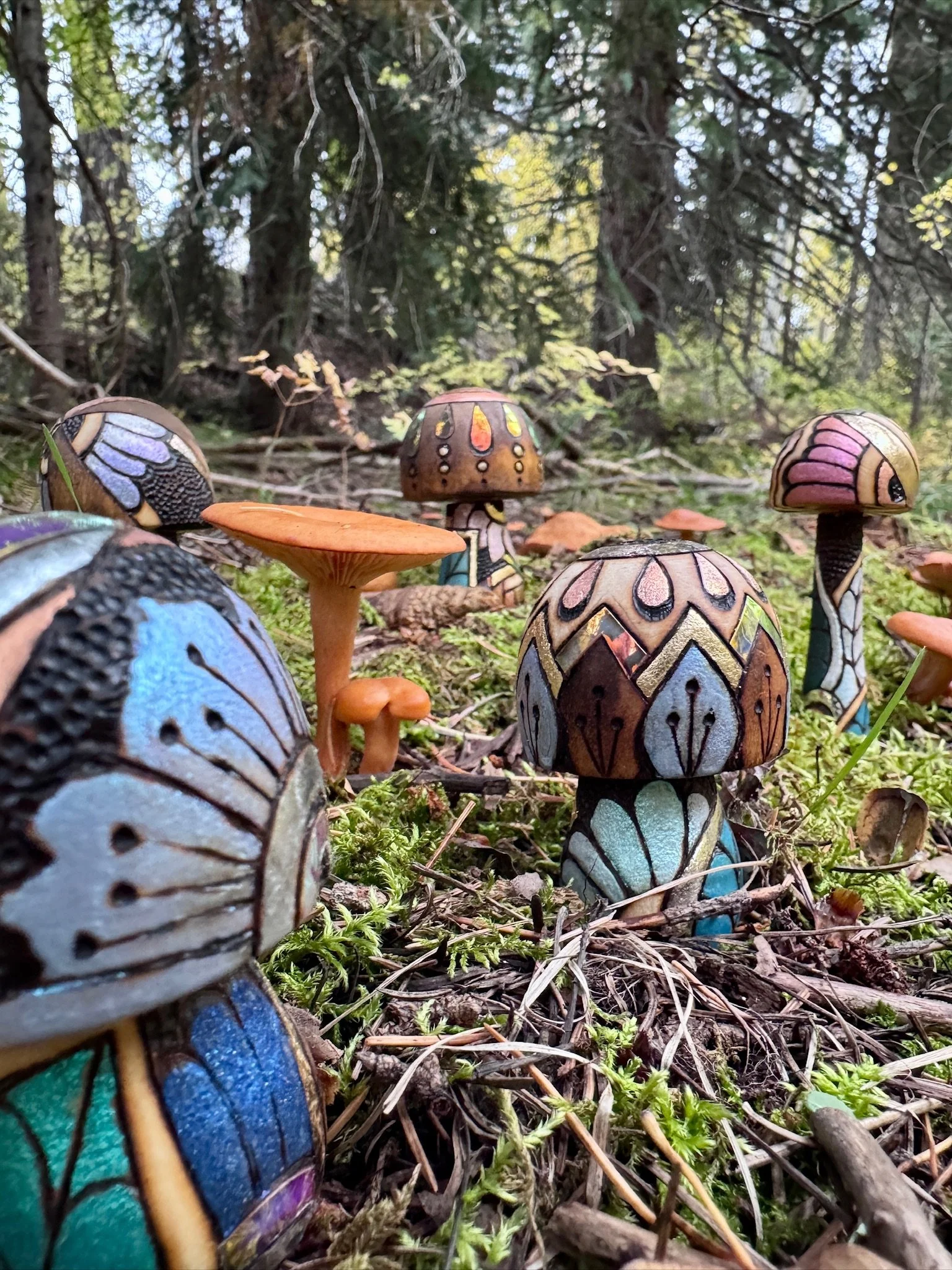Several handcrafted wooden mushrooms with colorful gilded patterns and light-reflective details arranged among natural woodland mushrooms.
