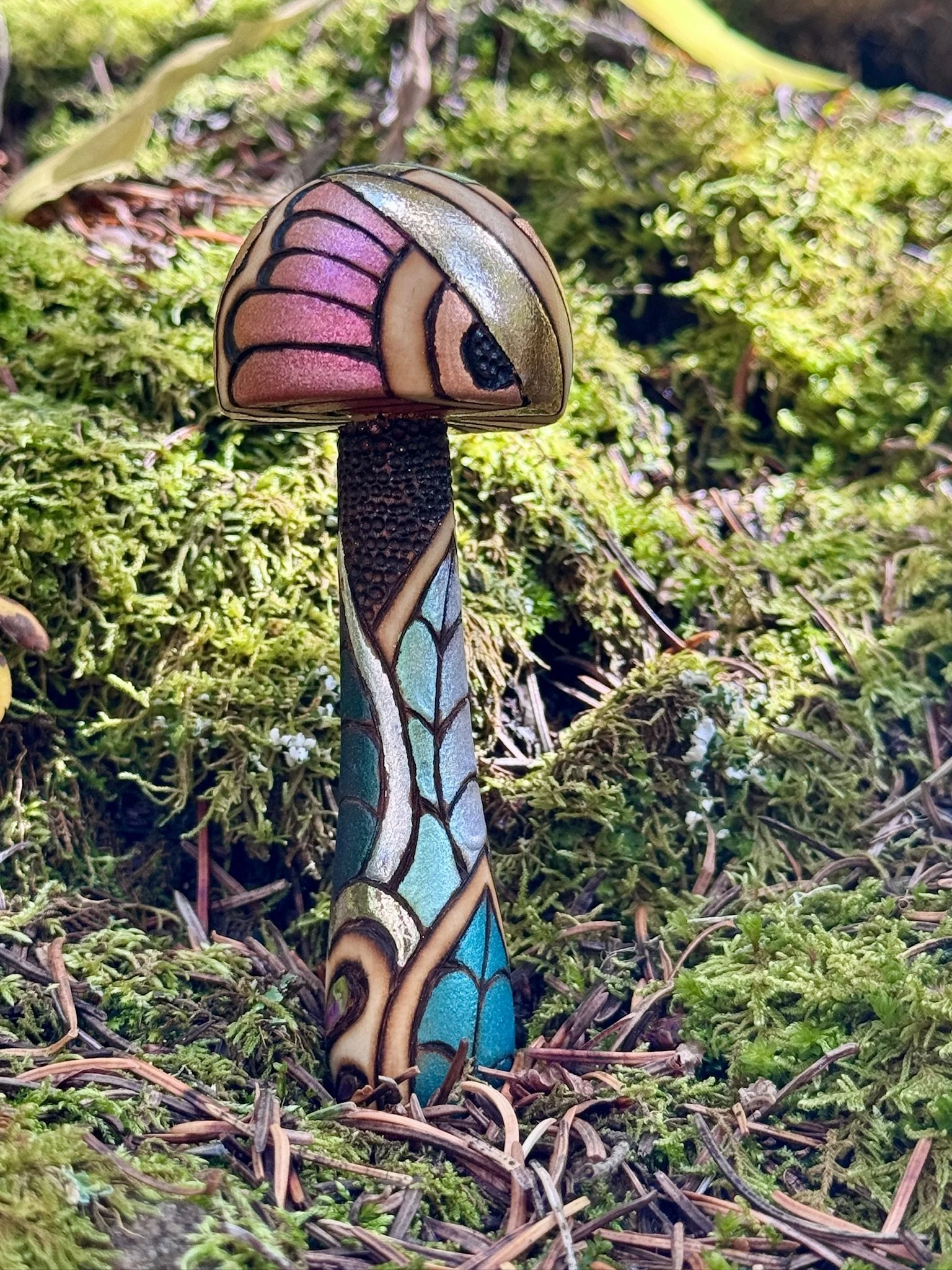 A single handmade wooden mushroom sculpture with a pink patterned cap, gold accents, and a turquoise botanical stem standing in green moss.