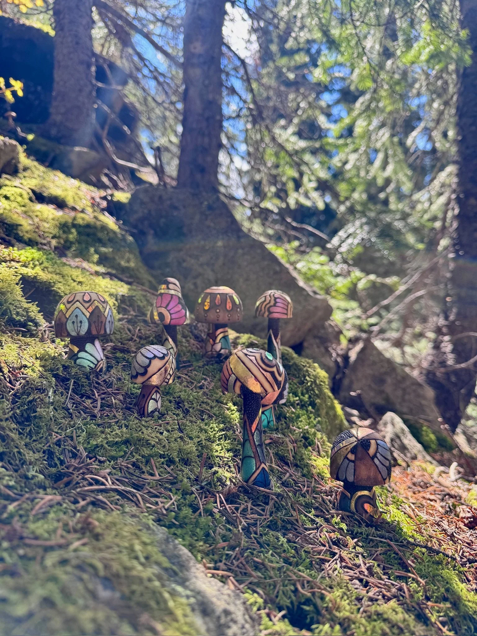A collection of handcrafted, light-responsive wooden mushroom sculptures with colorful gilded patterns arranged across a moss-covered forest hillside.