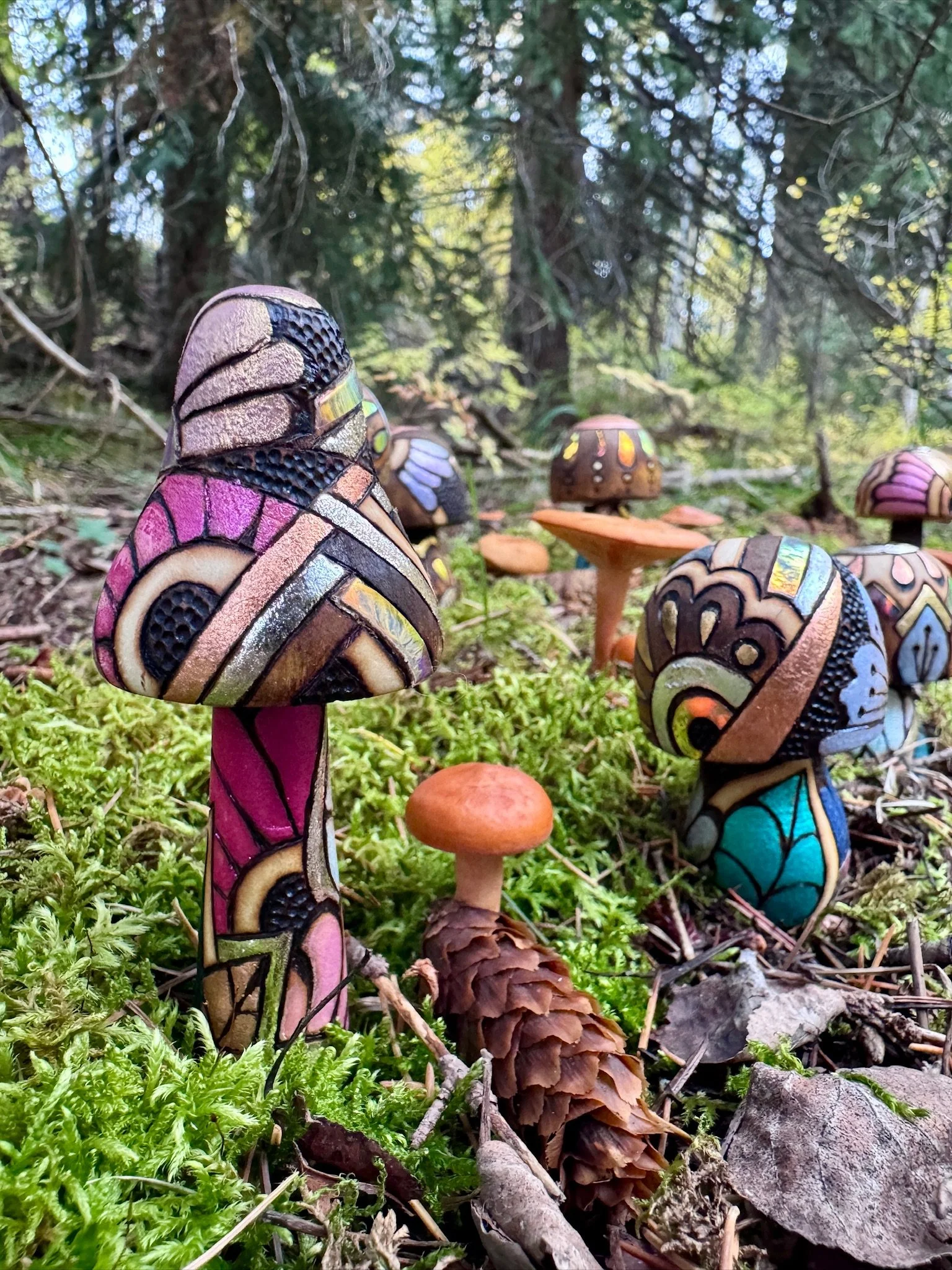 Colorful handcrafted wooden Firelight Shrooms by Austin artist Anne Shackelford, arranged among moss and pine needles with shimmering botanical patterns.