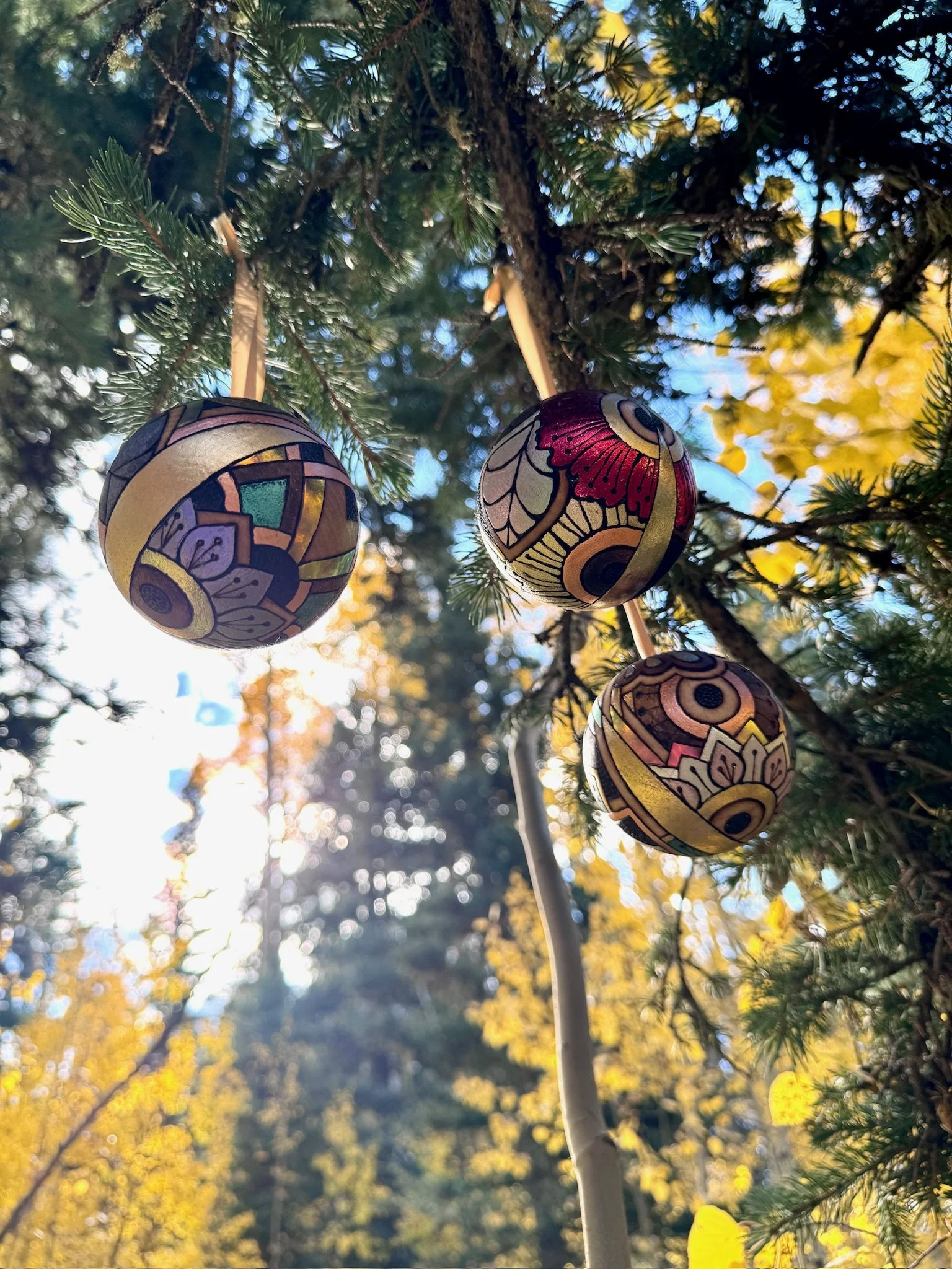 Three handcrafted Firelight Orbs hanging from tree branches, with colorful reflective patterns catching sunlight against golden autumn foliage.