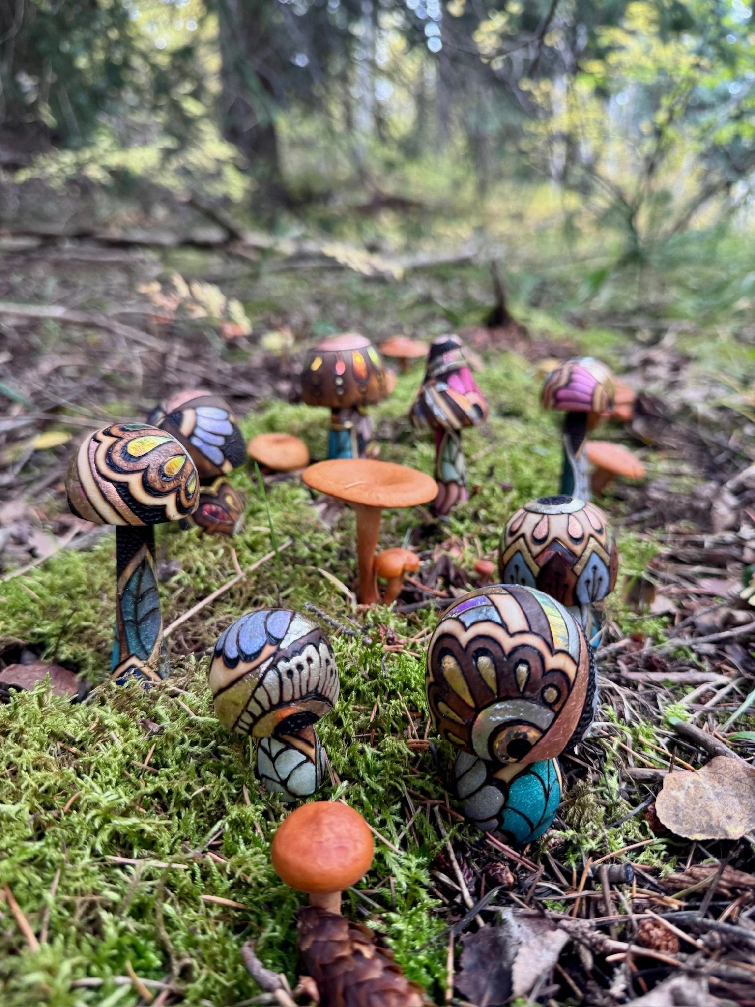 Handcrafted wooden mushrooms with blue, gold, and pastel botanical patterns surrounded by moss, pine needles, and wild mushrooms.