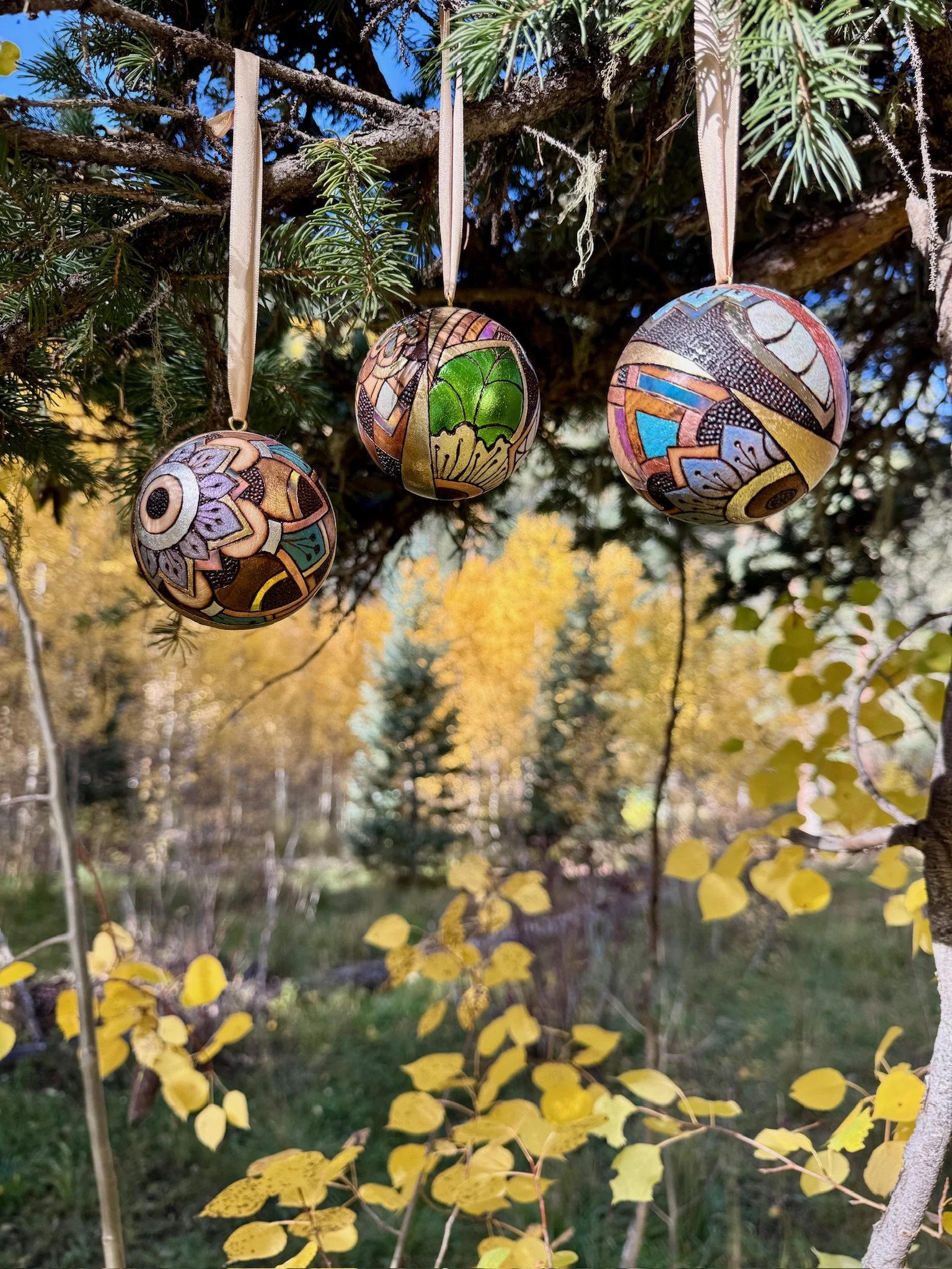 Three light-responsive wooden Firelight Orbs by Austin artist Anne Shackelford, suspended among evergreen branches and golden leaves.