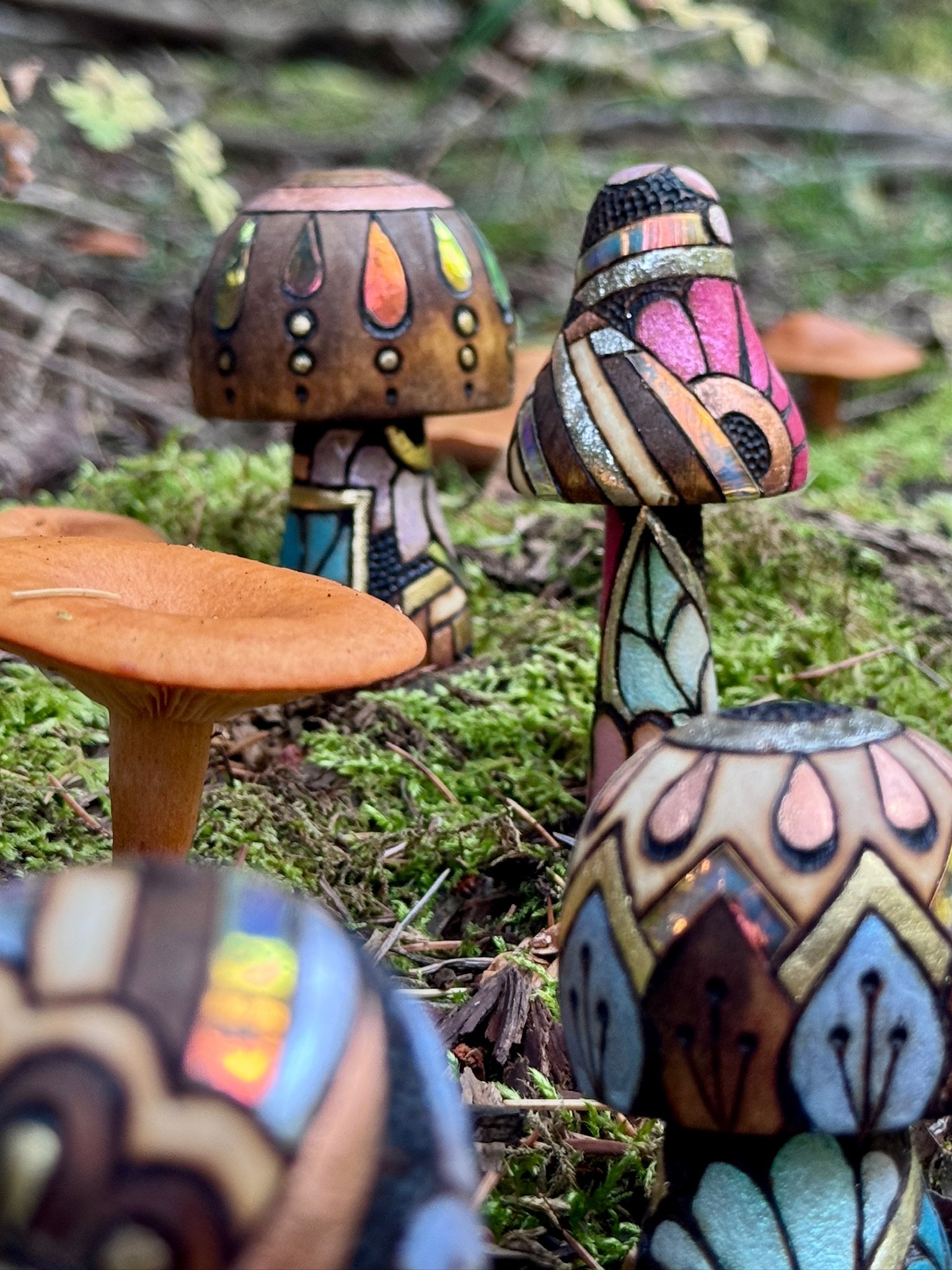 A collection of handmade wooden mushroom sculptures with colorful, reflective designs nestled in a moss-covered woodland setting.