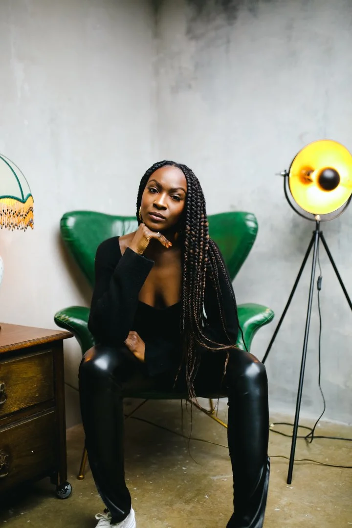 Portrait of a Black woman with long braided hair sitting in a green chair, wearing a black outfit, in a studio with a tripod lamp beside her.