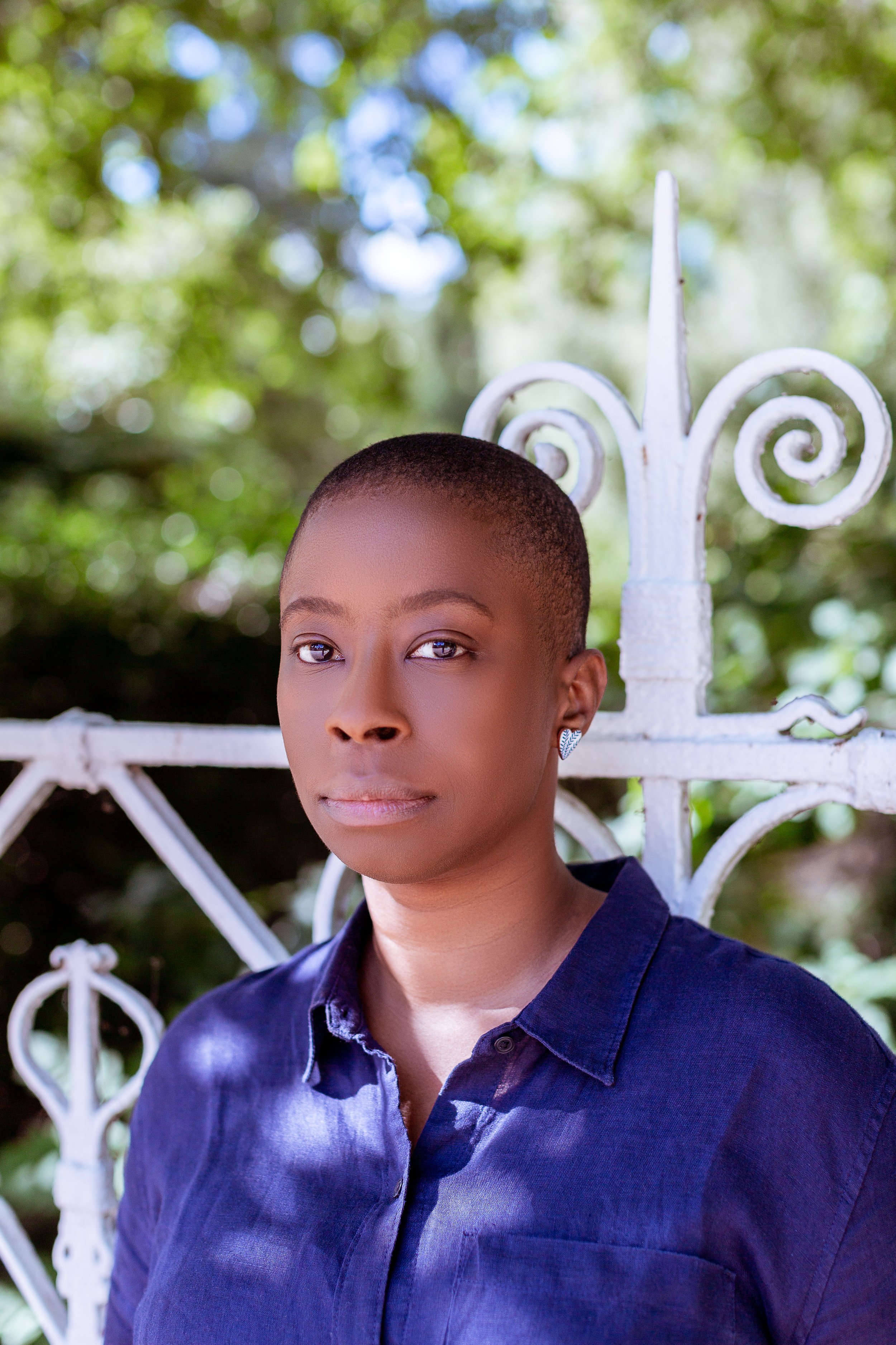 Portrait of a Black woman with a very short haircut wearing a dark blue shirt, standing outdoors in front of a decorative white gate with greenery behind