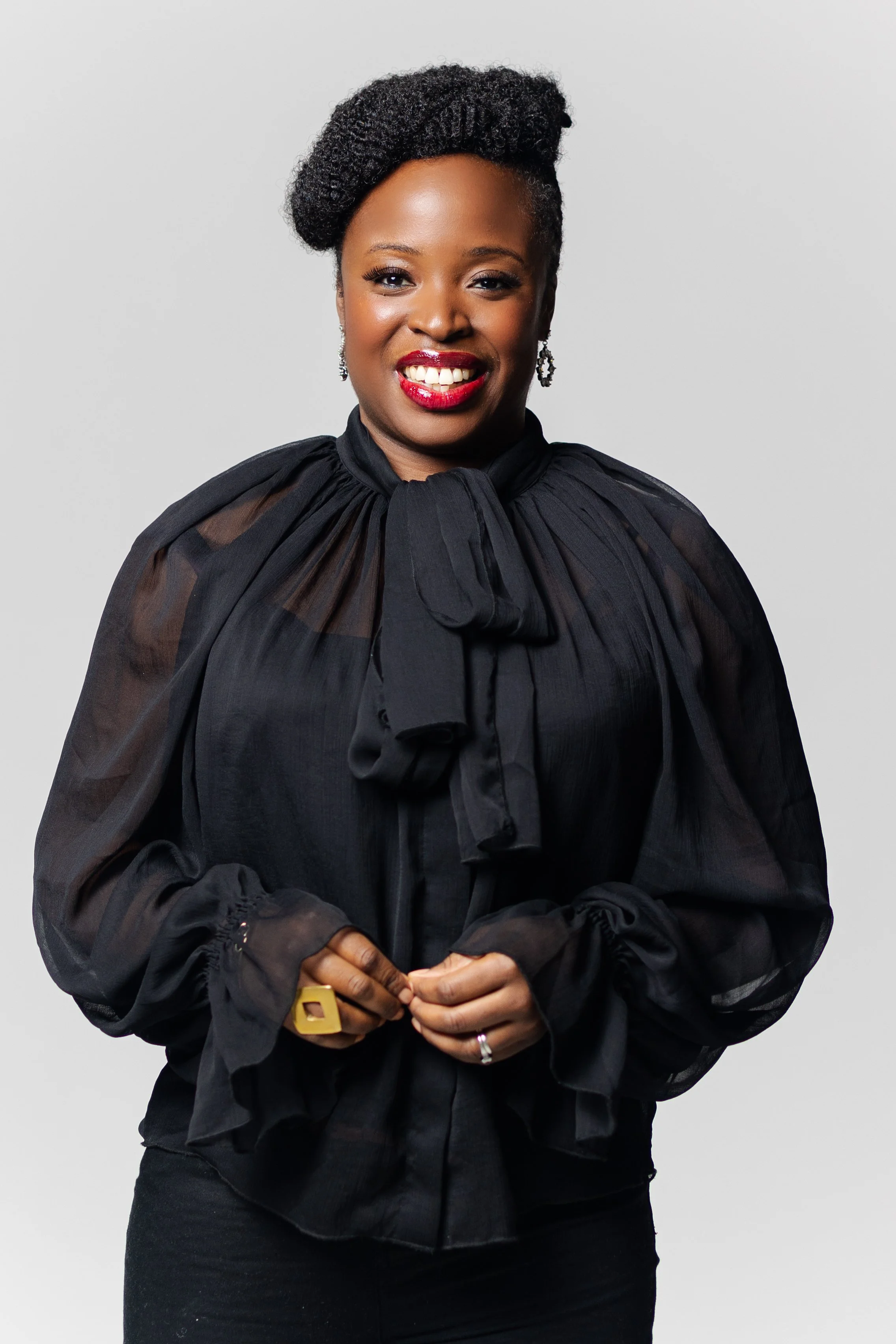 Close-up portrait of a smiling Black woman in a black sheer blouse, photographed against a plain light background.