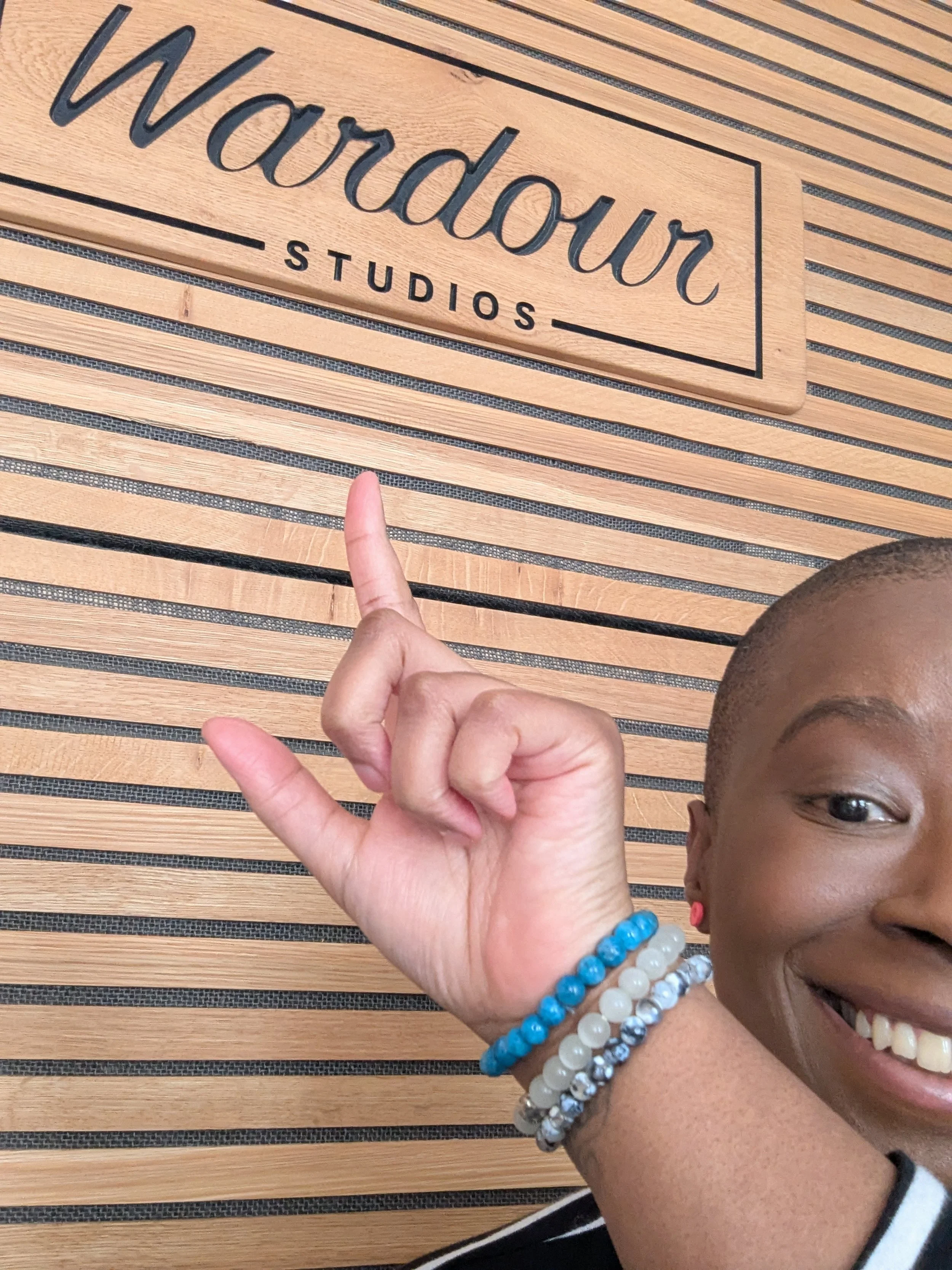 Cropped selfie of a smiling Black woman with very short hair pointing at a wooden wall sign that reads “Wardour Studios,” wearing beaded bracelets