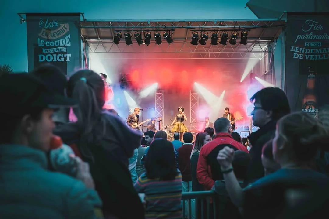 Scène d'un concert en plein air avec un groupe de musiciens sur scène, éclairages colorés, et un public regardant le spectacle.